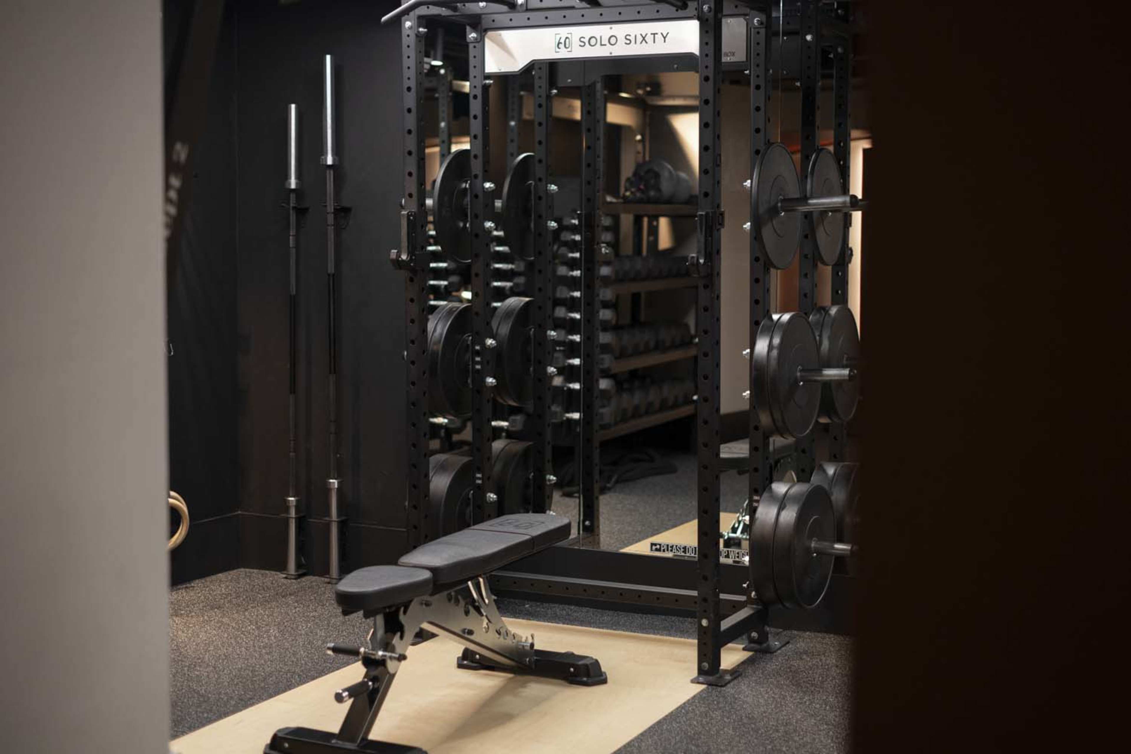 The image shows a gym space featuring a weightlifting rack with plates, a bench, and organized free weights on shelves.