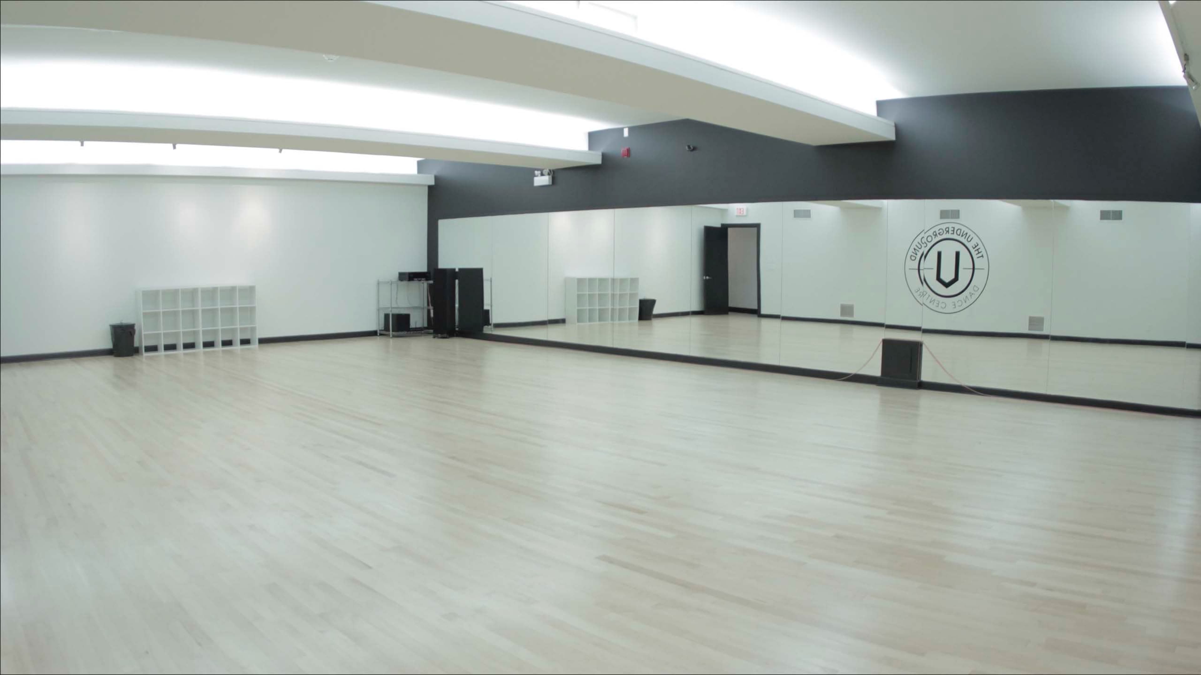 A spacious, empty dance studio with wooden flooring, large mirrors on one wall, and minimal storage units.