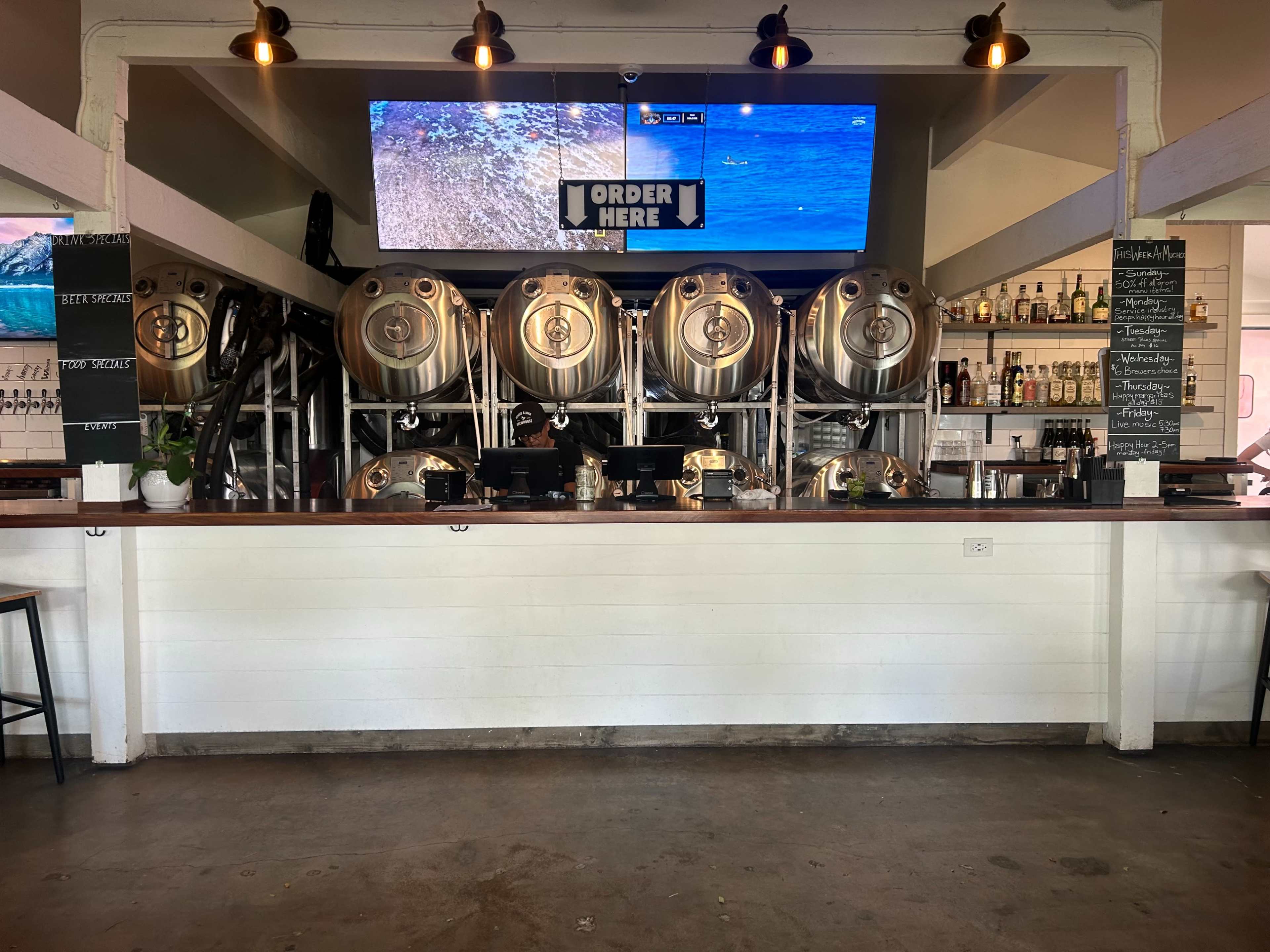 The image shows a modern bar counter with large metal brewing tanks behind it, and two screens displaying visuals above.