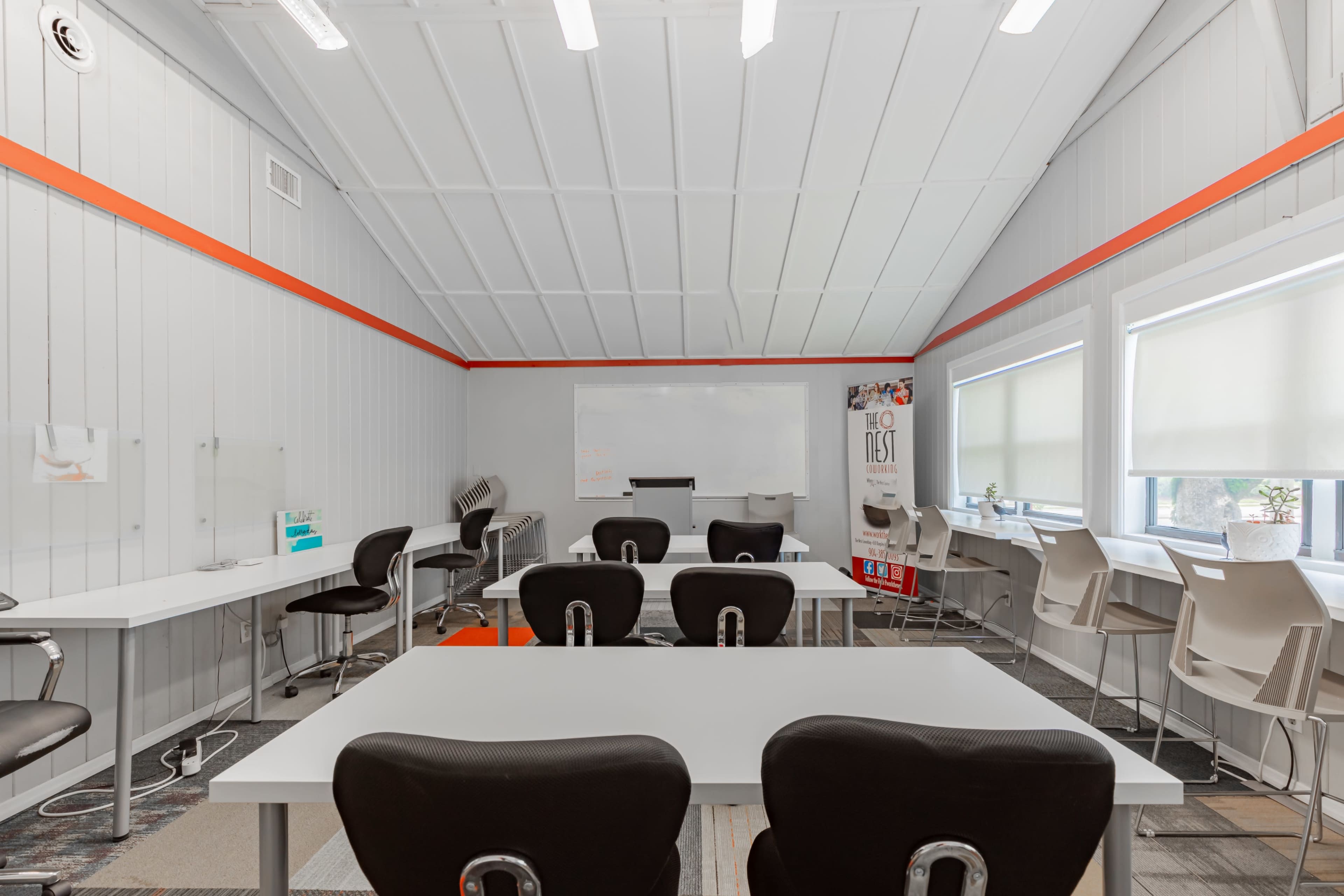 A modern classroom features white tables and black chairs arranged for group work, with large windows allowing natural light and a whiteboard at the front.
