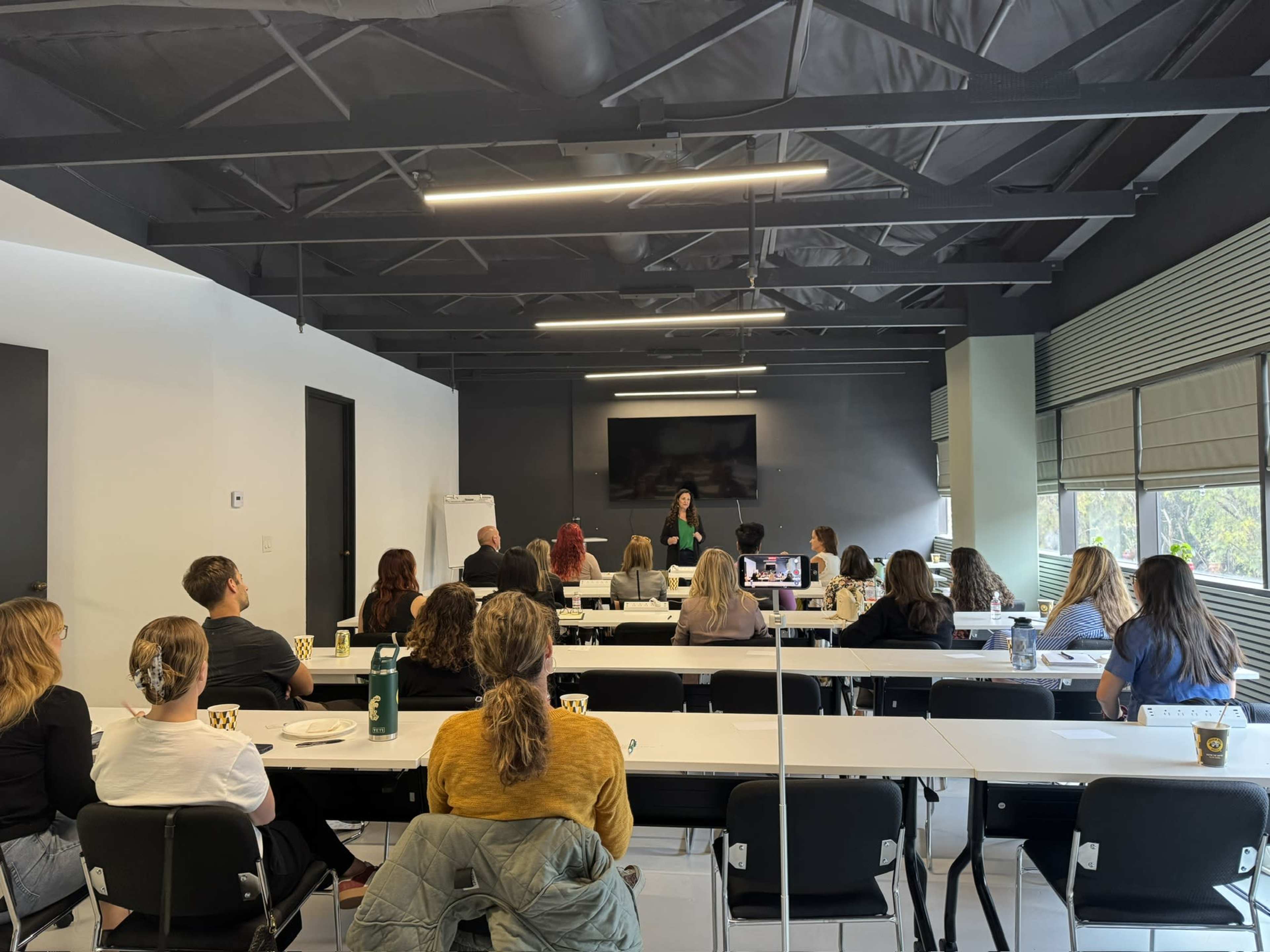 A group of individuals sits in a conference room, attentively listening to a speaker at the front of the room.