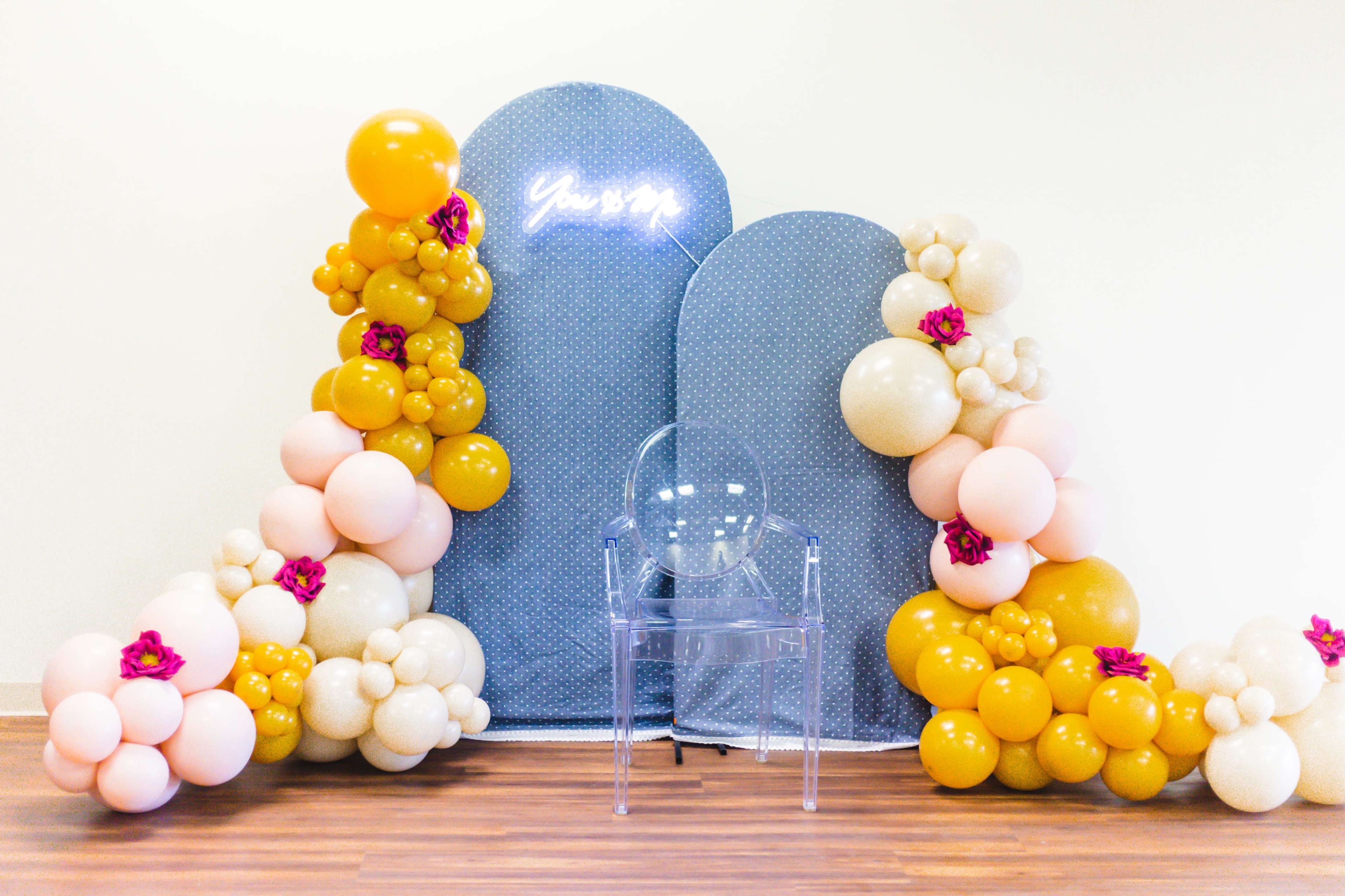 A decorative backdrop with large, round shapes adorned with colorful balloons and a clear acrylic chair in front of it.