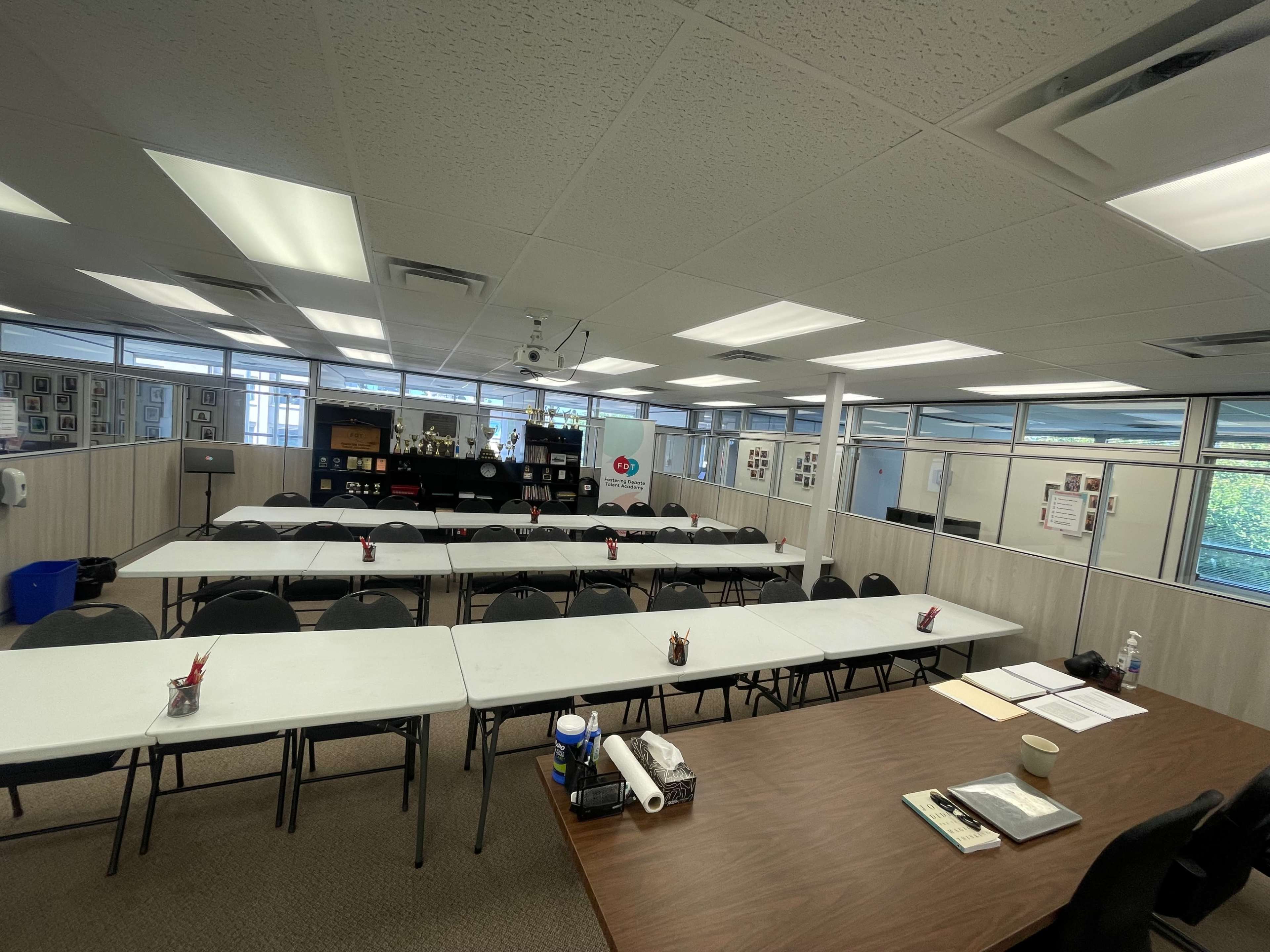 A large meeting room with several tables and chairs arranged for a gathering, featuring windows along the walls and various materials on the tables.