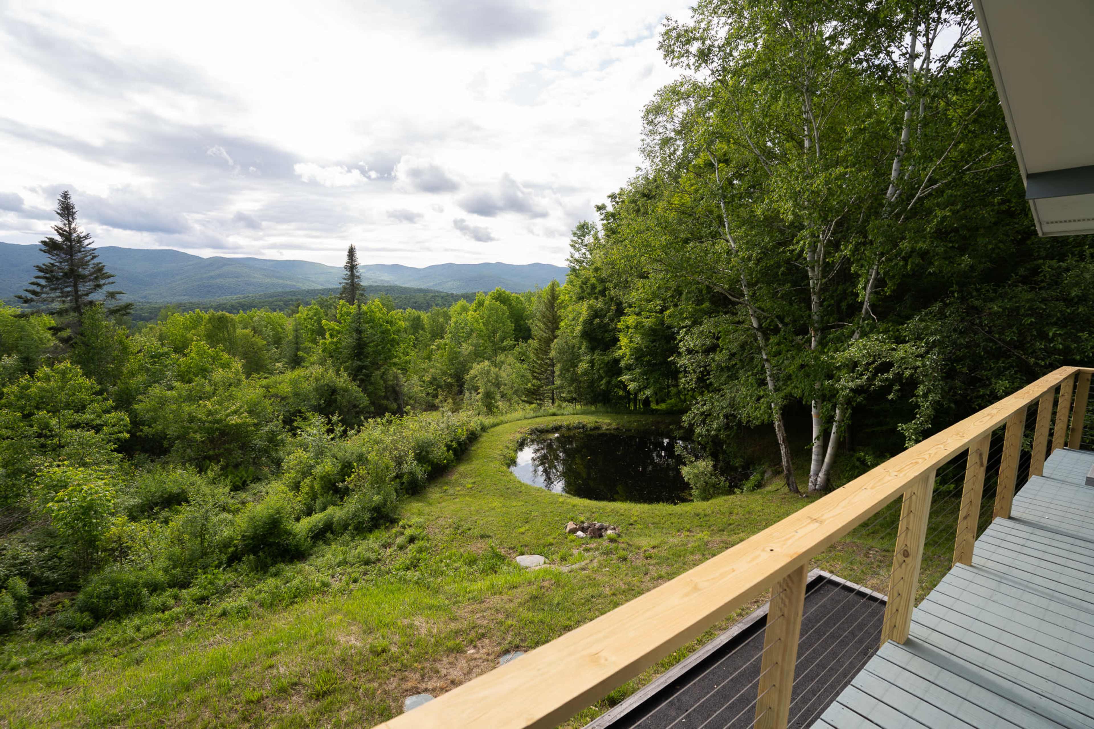 A wooden deck overlooking a pond surrounded by trees and mountains in the distance under a cloudy sky.