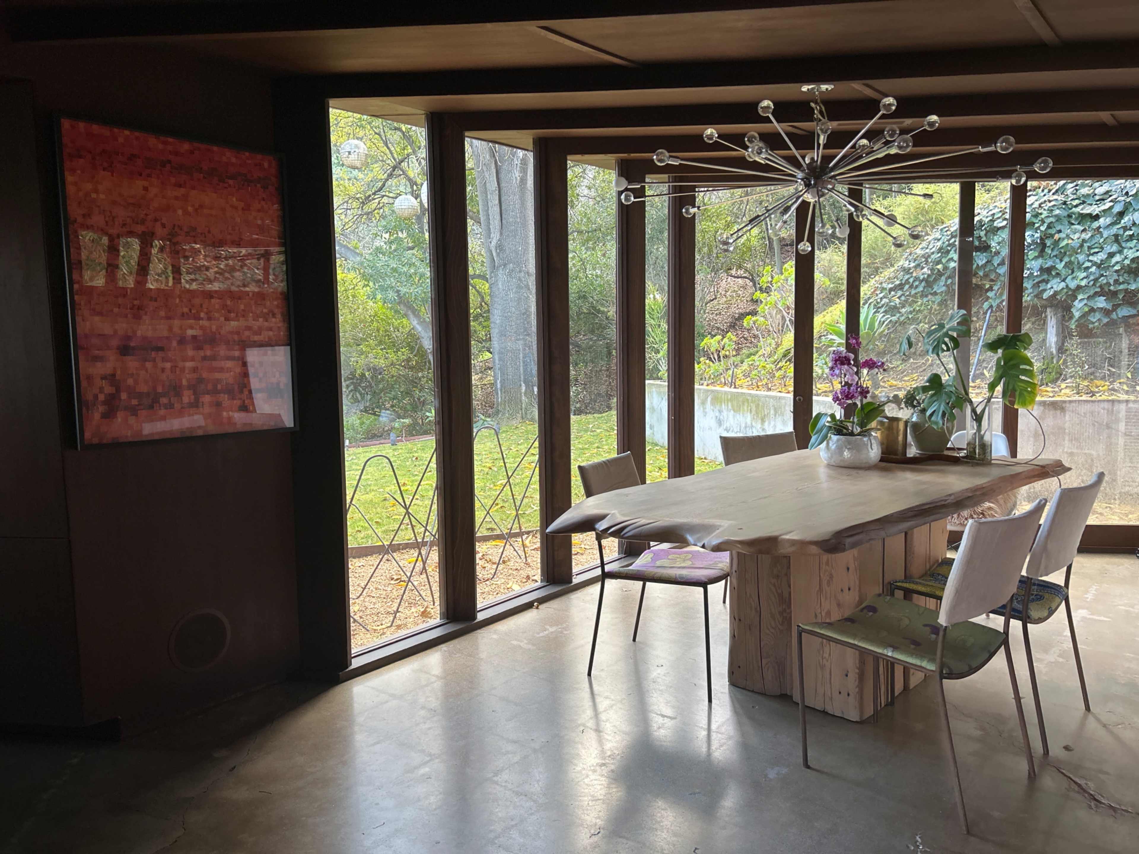 A dining area features a large wooden table surrounded by chairs, with large windows revealing a view of an outdoor garden.