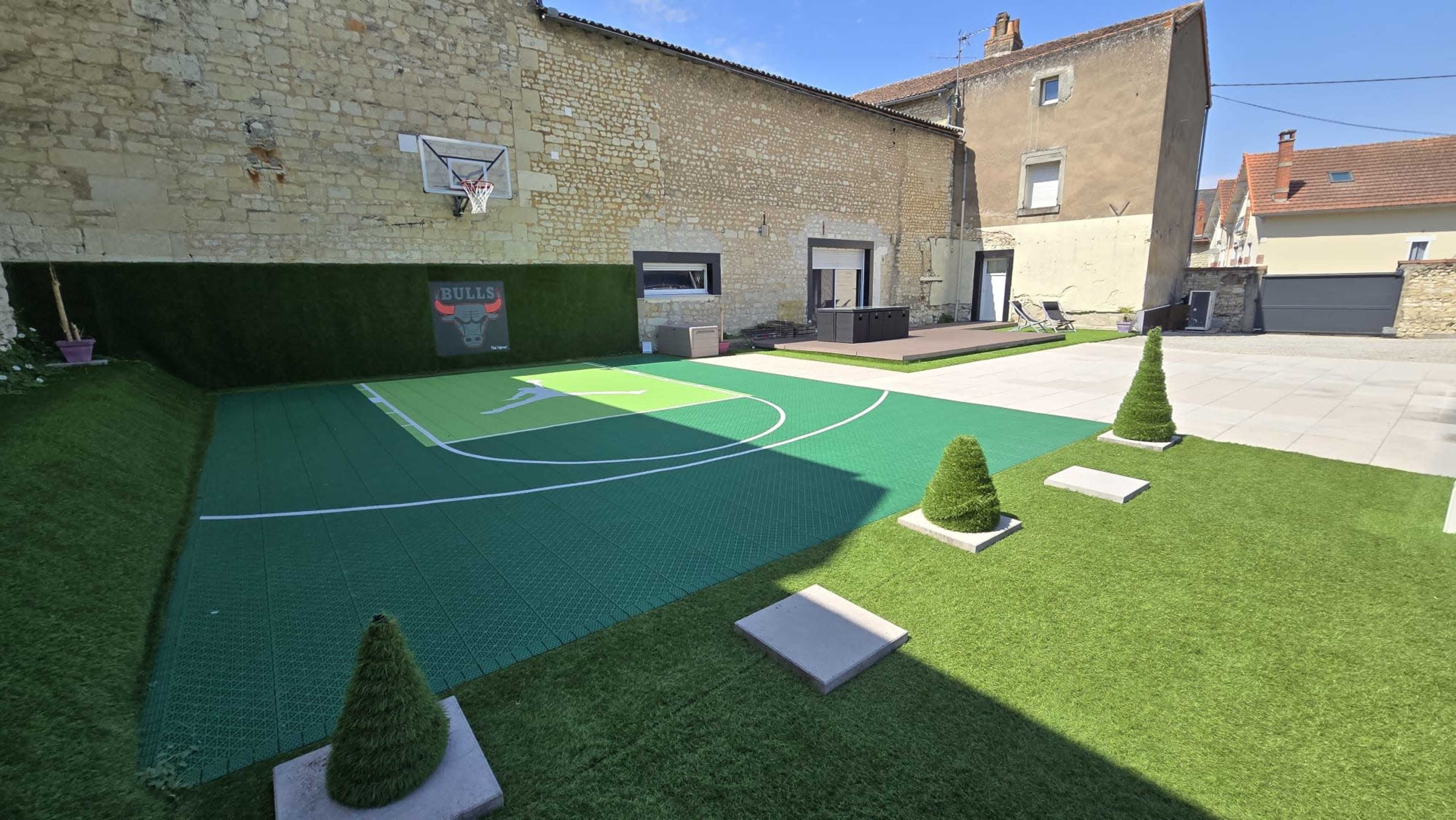 The image shows a small outdoor basketball court with a green turf surface, surrounded by decorative bushes, and a building in the background.