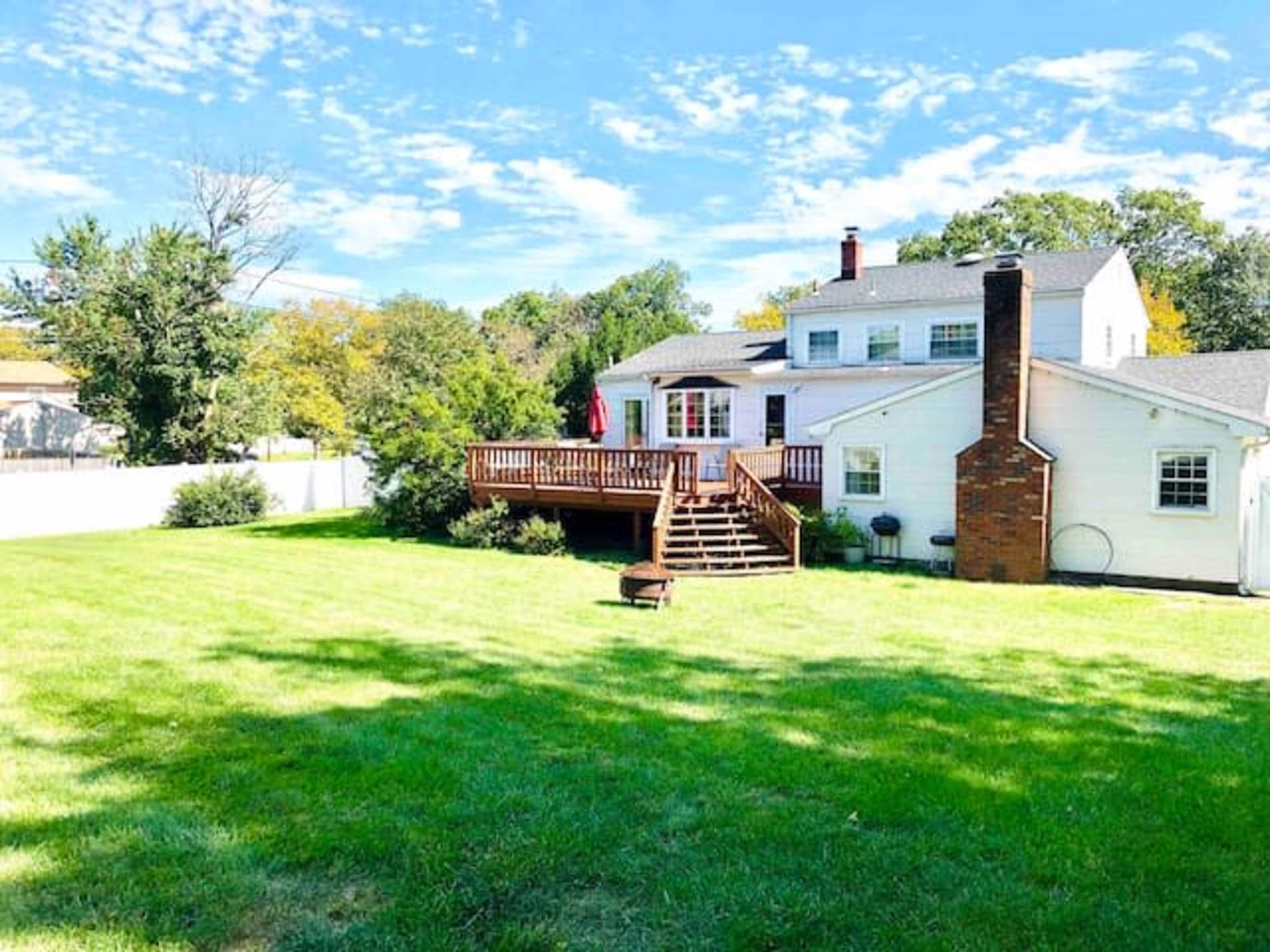 A backyard with a large green lawn, a wooden deck attached to a two-story white house, and a brick chimney.