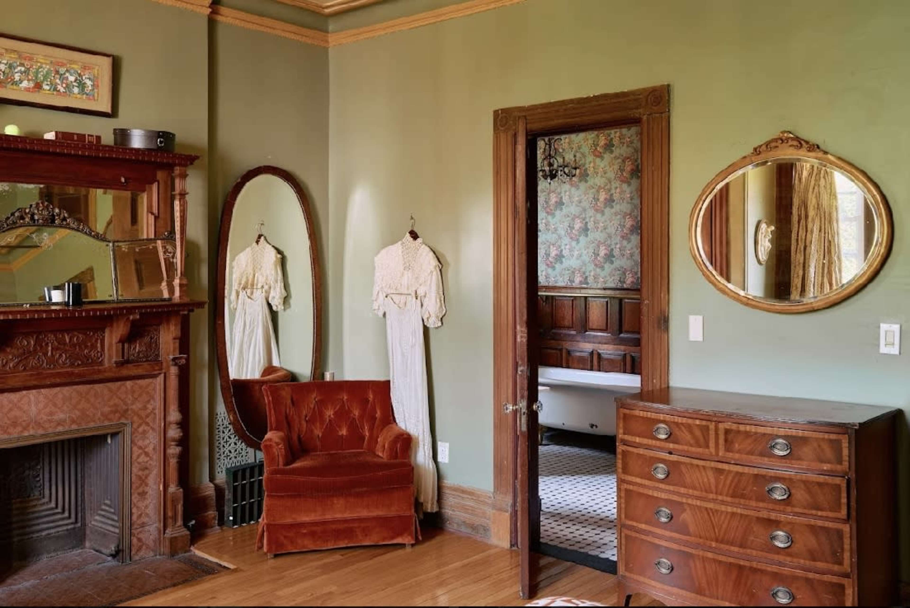 The image shows a vintage-style room featuring a red velvet armchair, a wooden dresser, a large mirror, and a door leading to another area, with floral wallpaper and two hanging garments.