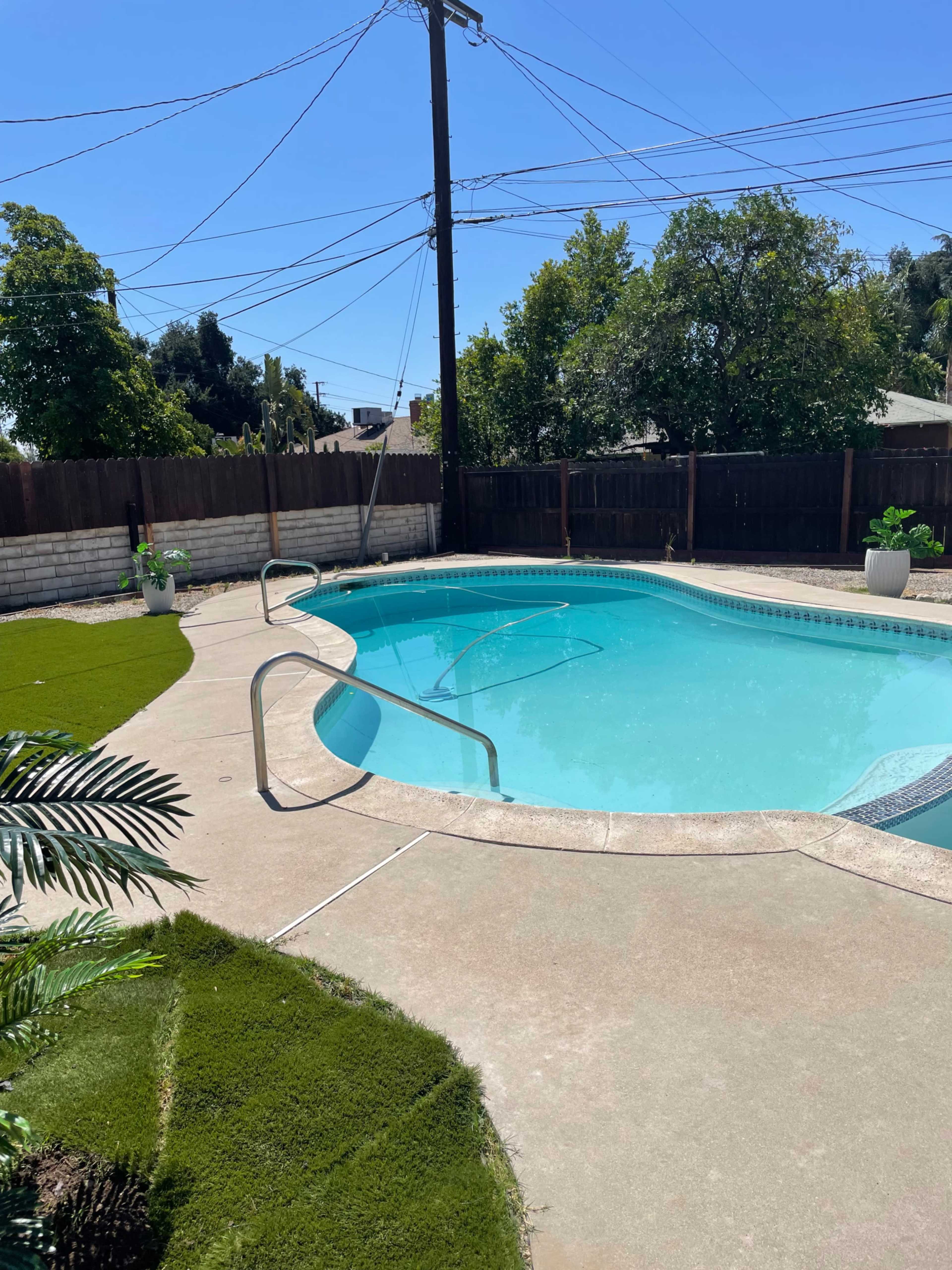 The image shows a clear swimming pool surrounded by a concrete deck and greenery under a bright blue sky.