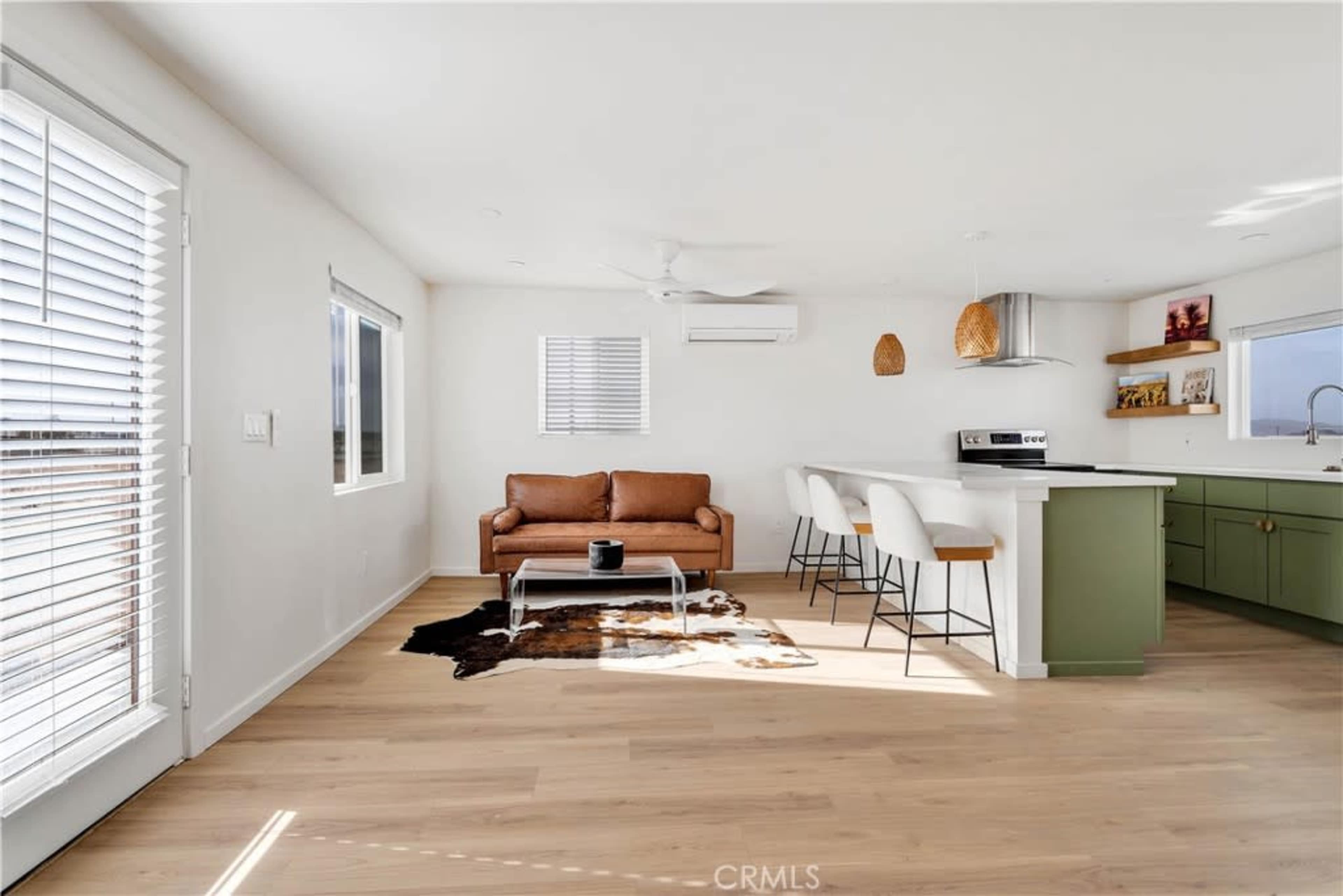 A bright, modern living space featuring a brown leather sofa, a kitchen island with bar stools, and large windows that provide natural light.