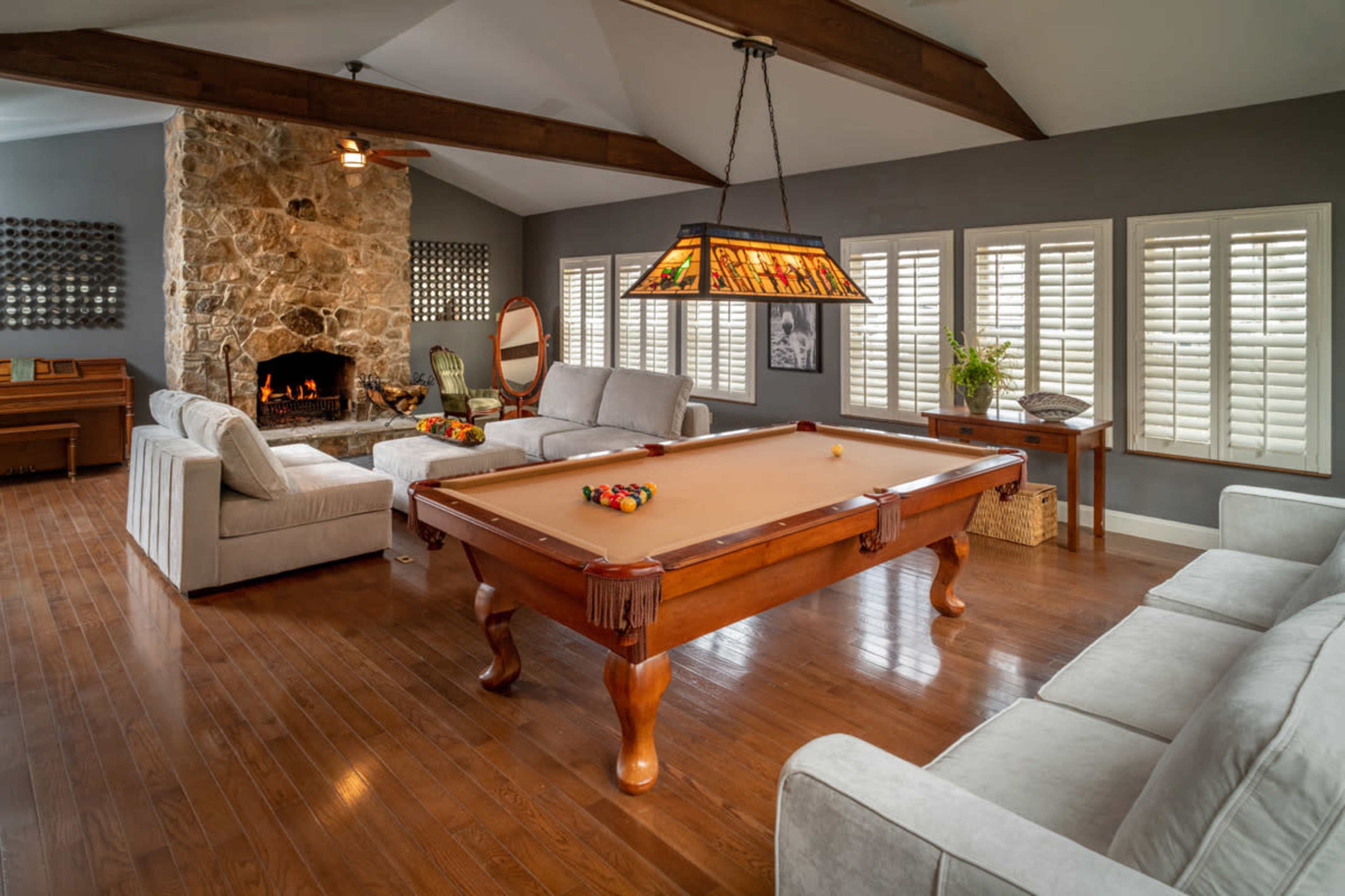 The image shows a living room with a slate pool table at the center, surrounded by two sofas, a stone fireplace in the background, and large windows with shutters.