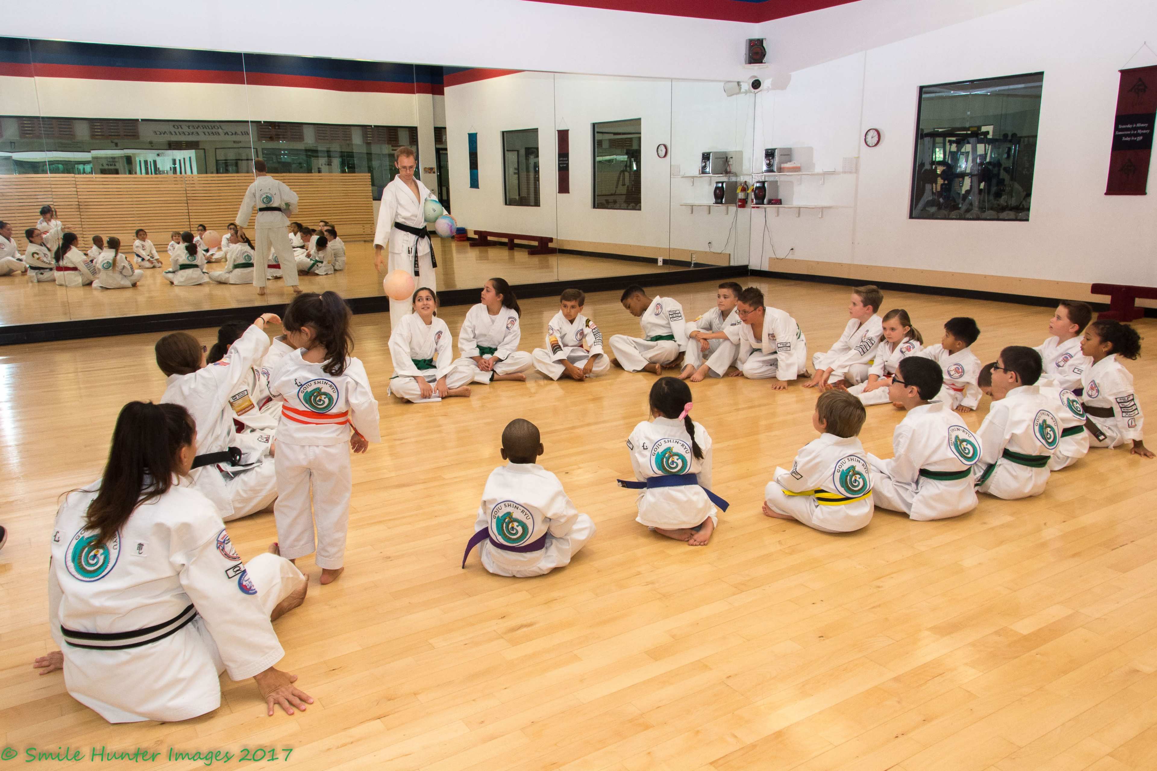 A group of children and instructors sits in a circle on the mat in a martial arts studio, with some participants engaging in activities while others watch.