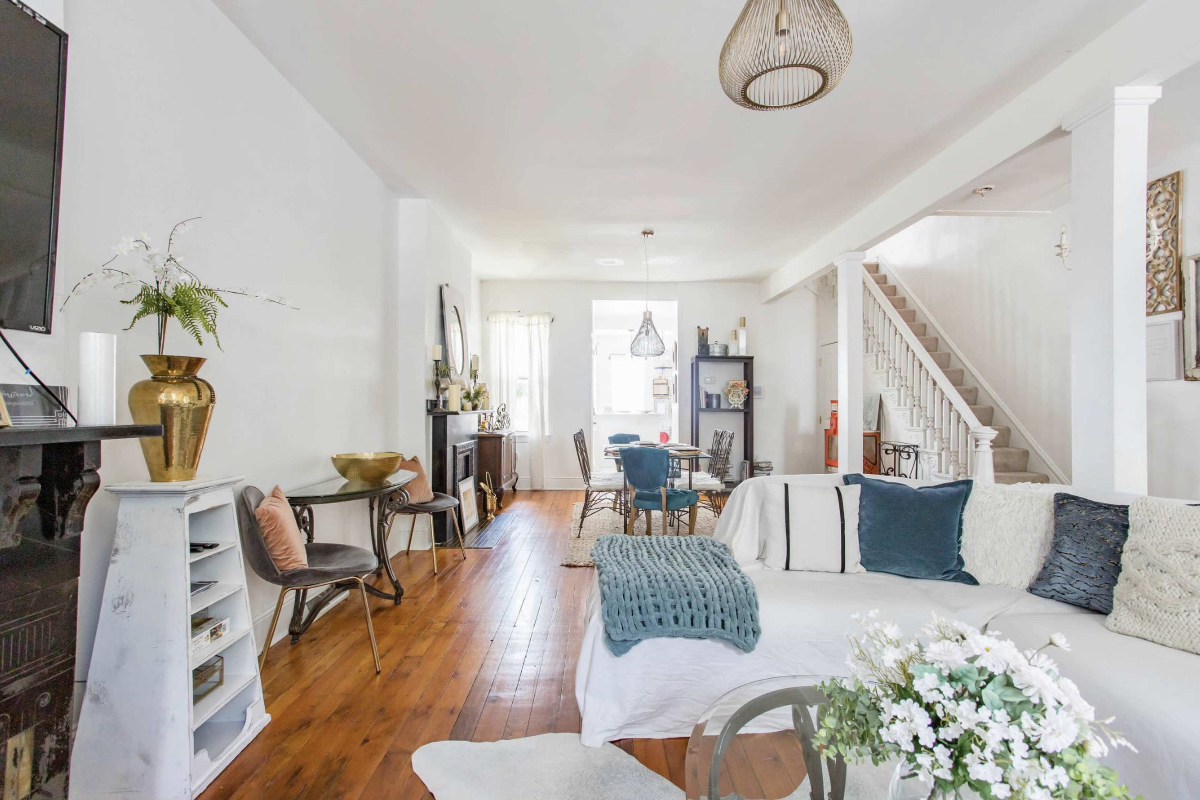 The living room features a white sofa, wooden flooring, and a staircase leading to an upper level, with various decorative elements and furniture throughout the space.