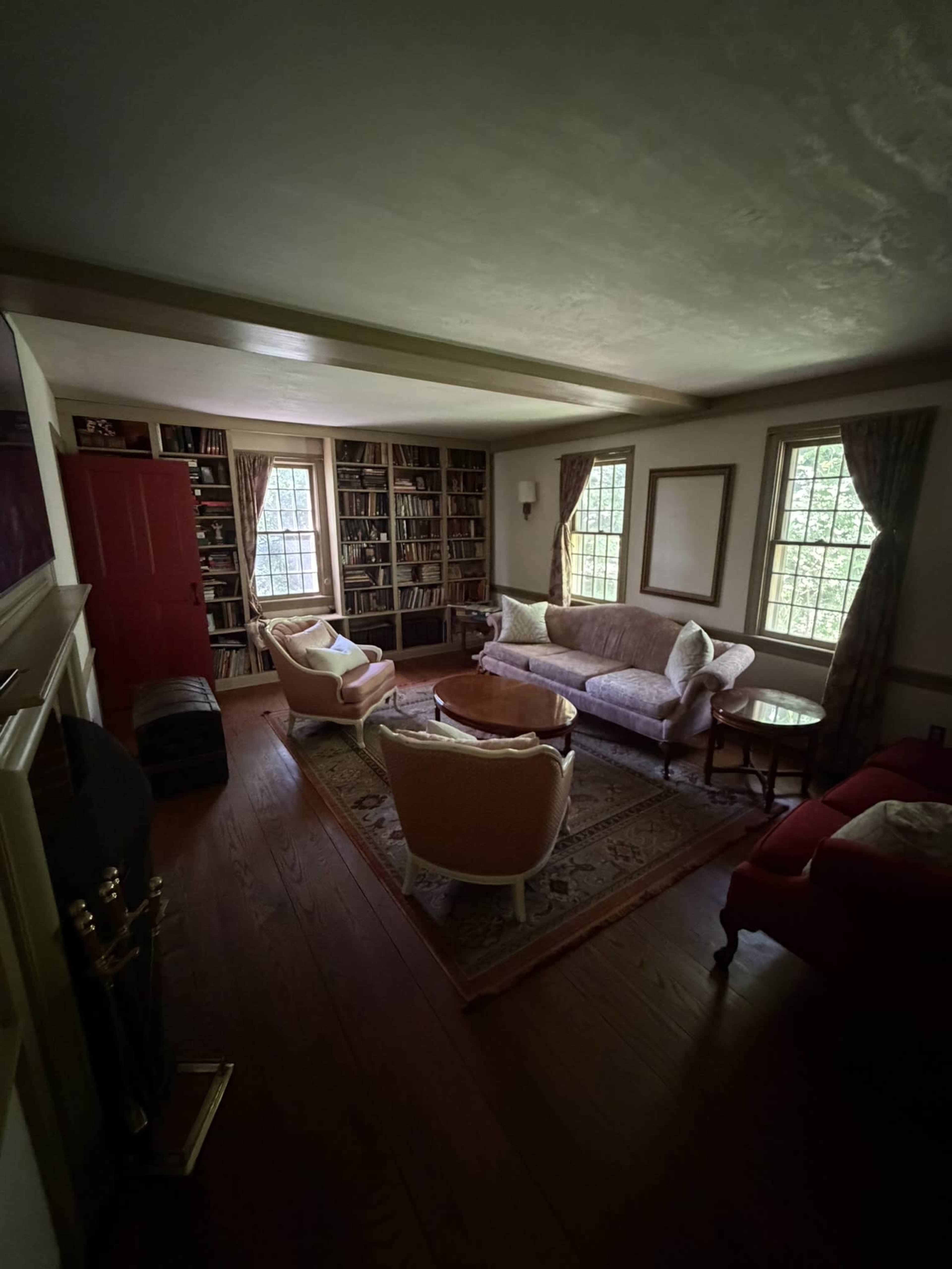 The image shows a cozy living room with bookshelves lining the walls, two armchairs, a sofa, and a wooden table near a window.