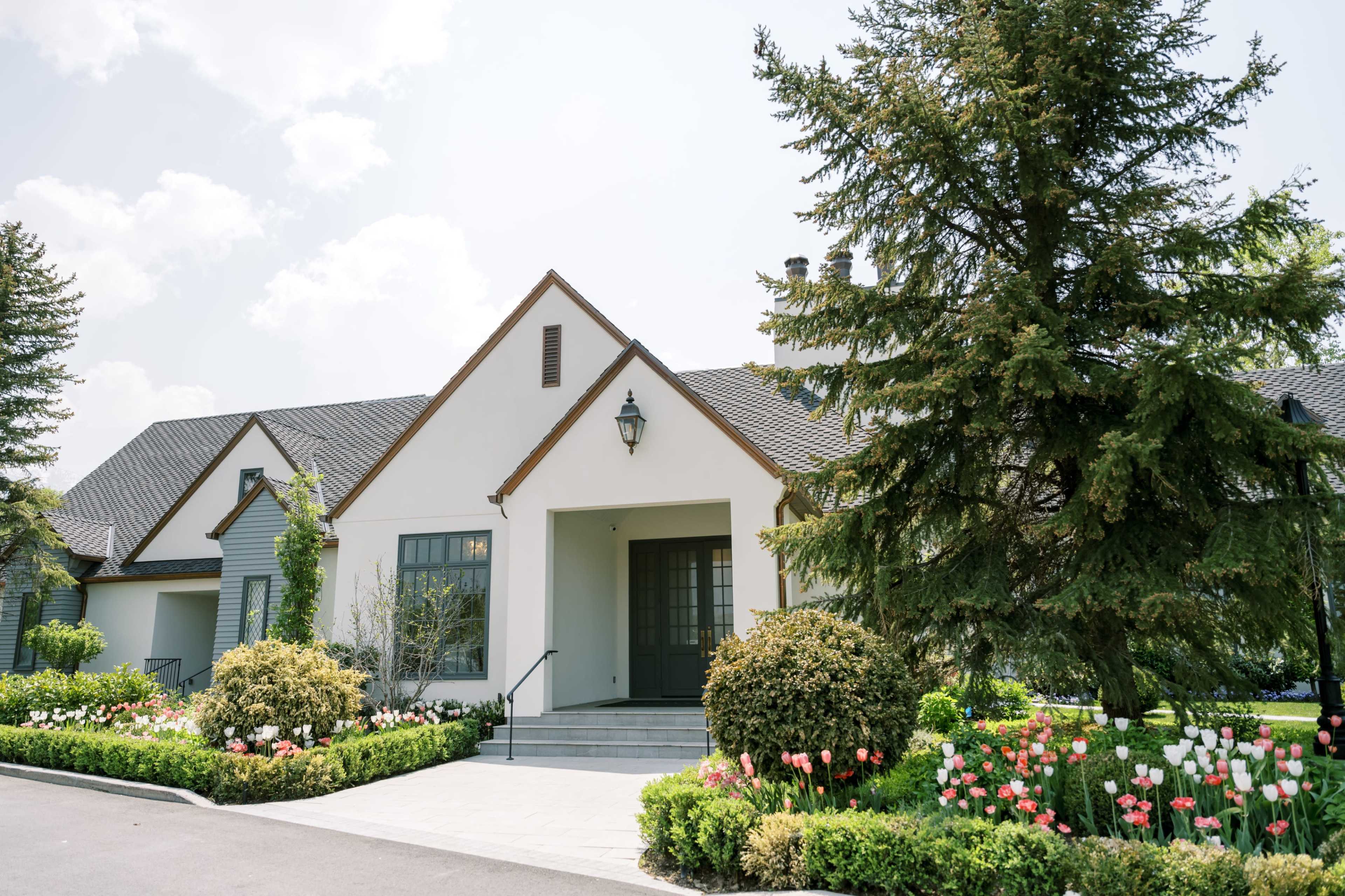 The image shows a modern house with a landscaped garden featuring colorful flower beds and a pathway leading to the entrance.