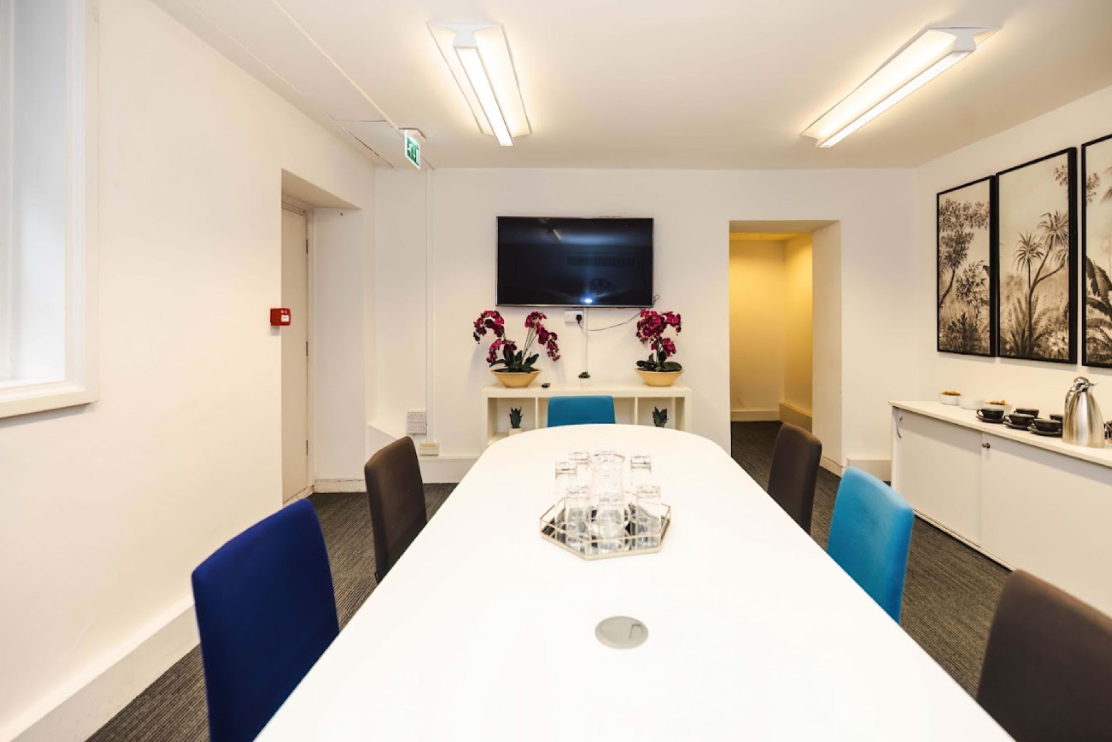 A modern meeting room features a white table surrounded by chairs, a large screen on the wall, and decorative plants.
