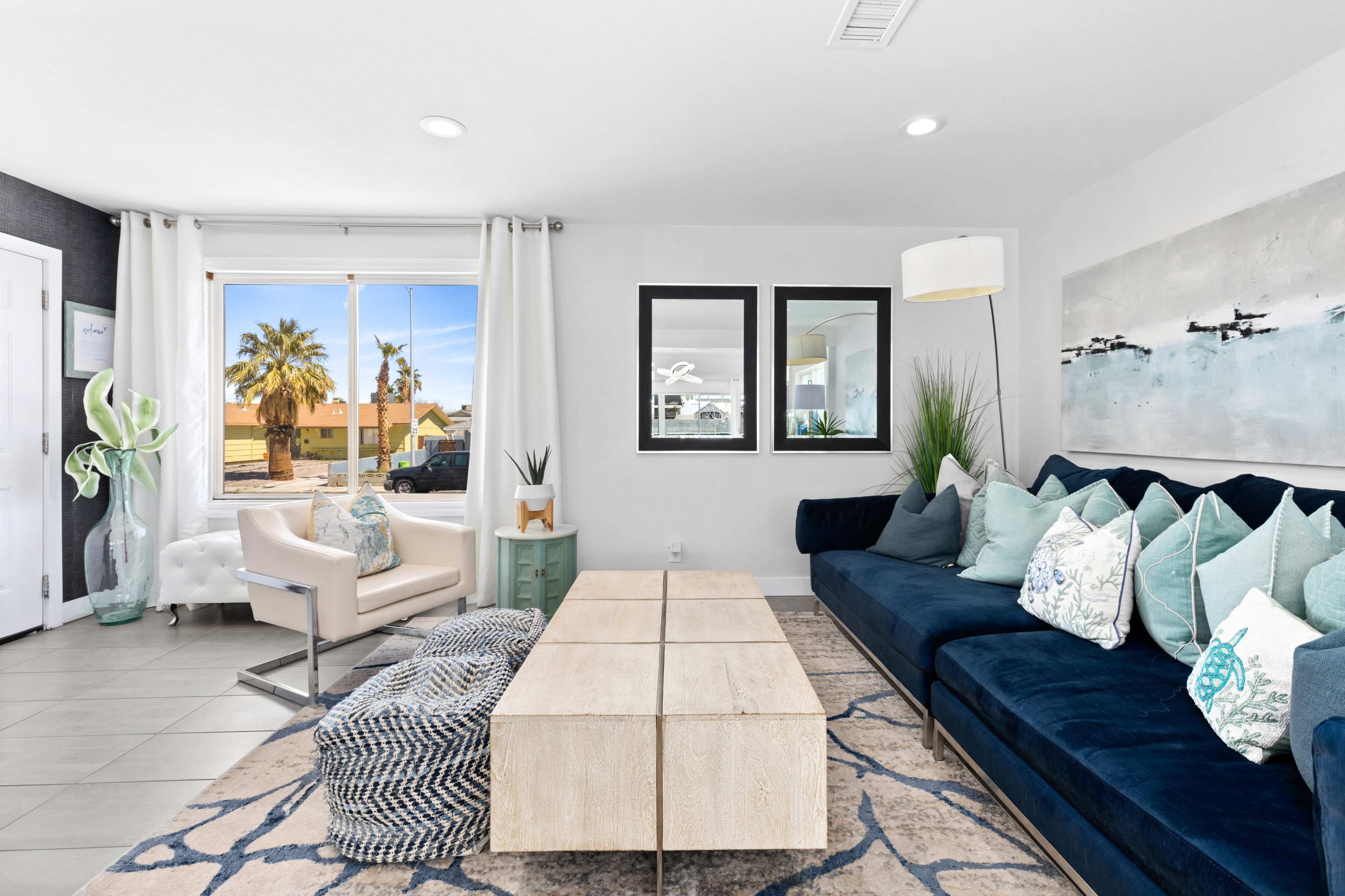 A modern living room features a dark blue couch adorned with various pillows, a light wooden coffee table, and large windows showcasing palm trees and outside scenery.