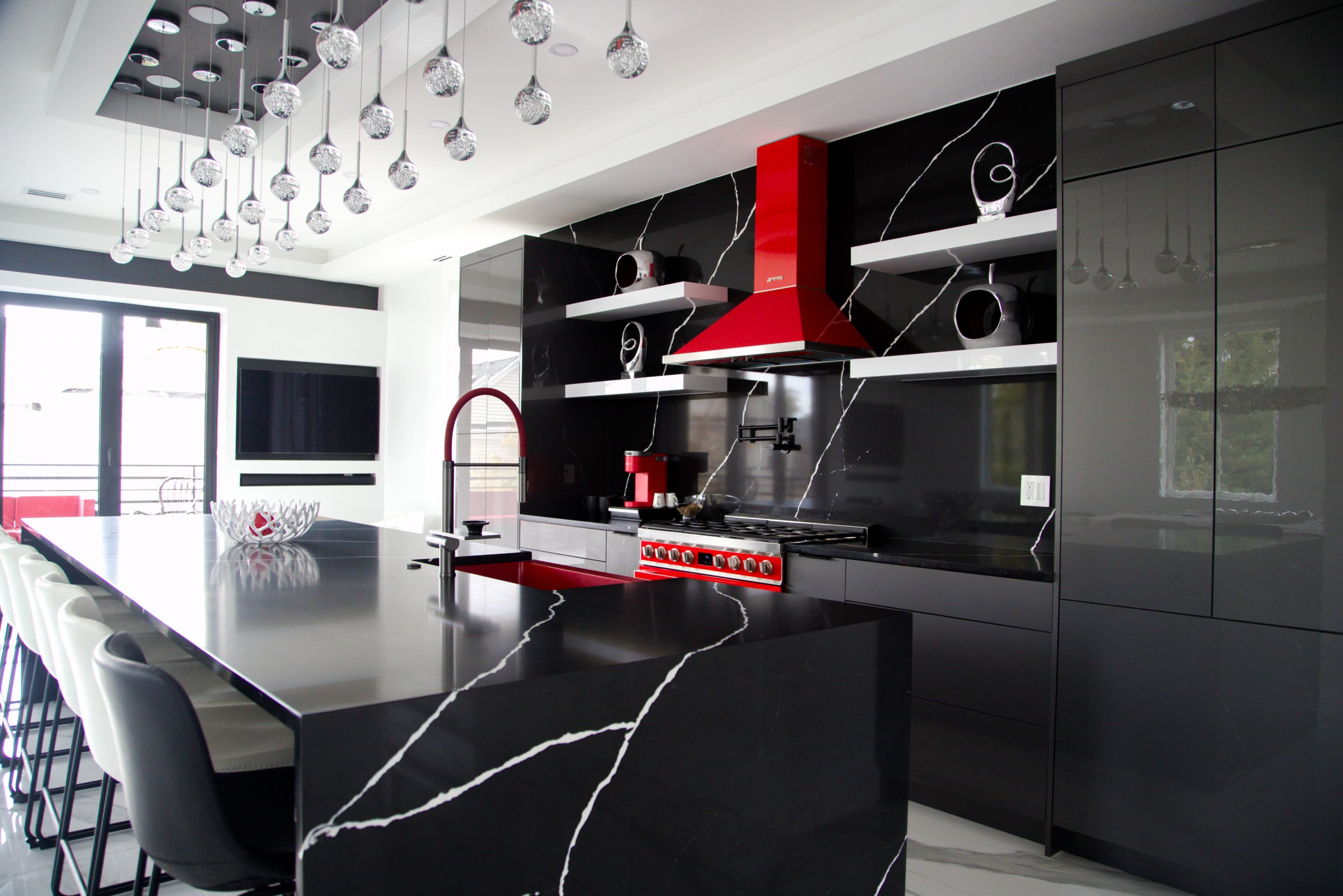The image shows a modern kitchen with a black and white color scheme, featuring a large island with seating, a red stove and hood, and decorative pendant lights overhead.
