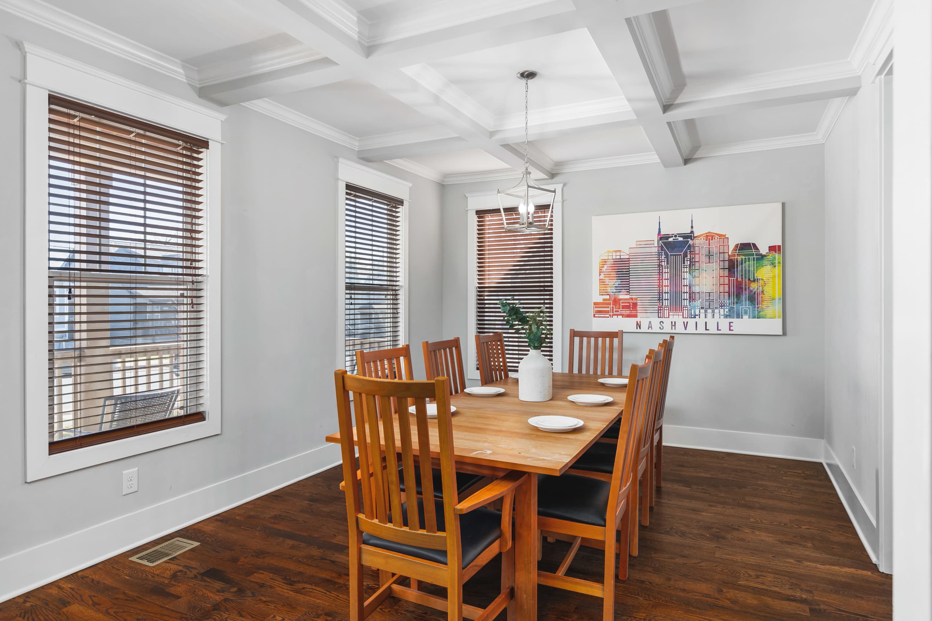 A dining room features a wooden table surrounded by eight chairs, with a colorful Nashville-themed artwork on the wall and natural light coming through the window blinds.