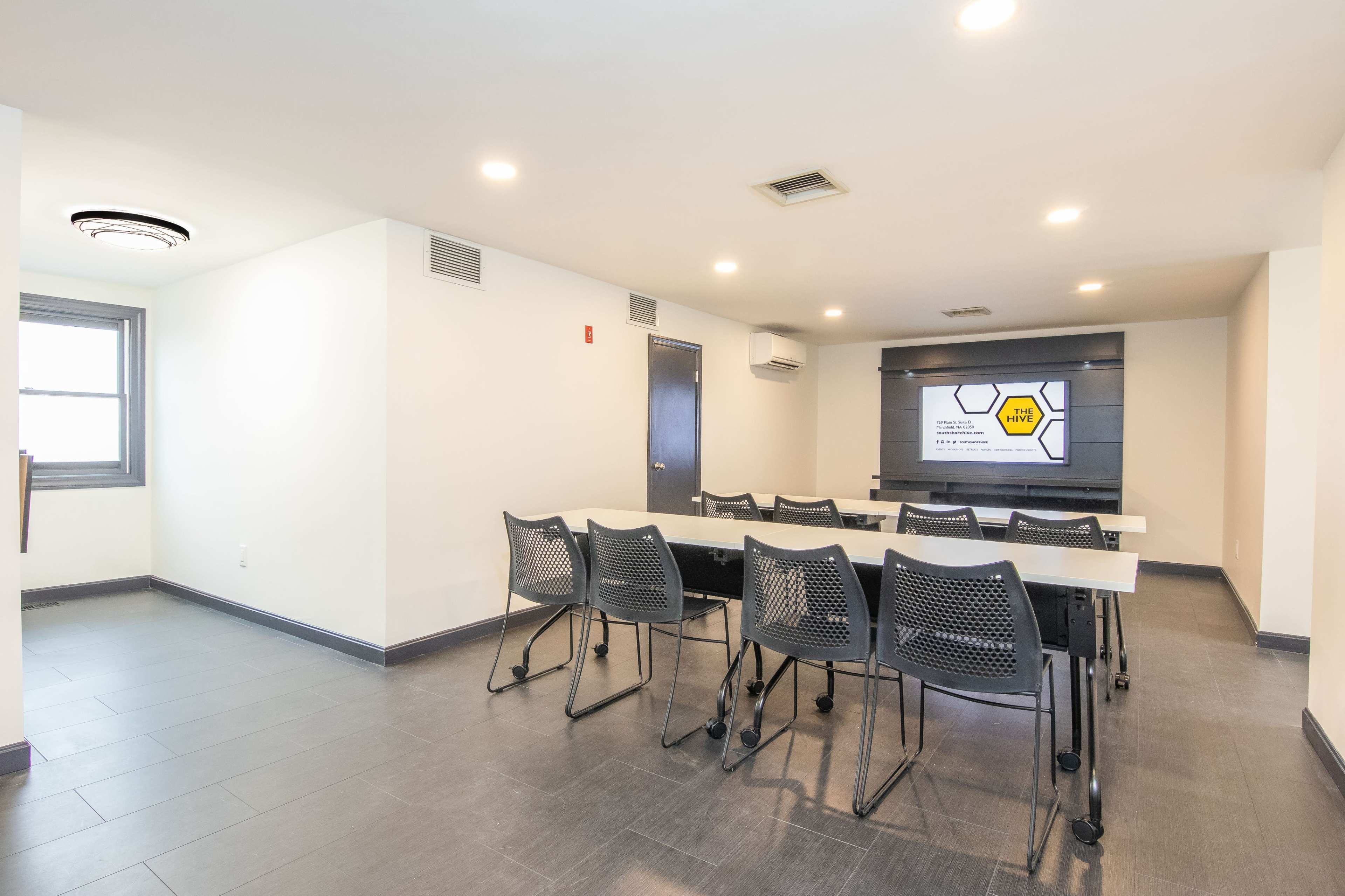 A conference room features a long table with rolling chairs and a large screen displaying a presentation on one wall.