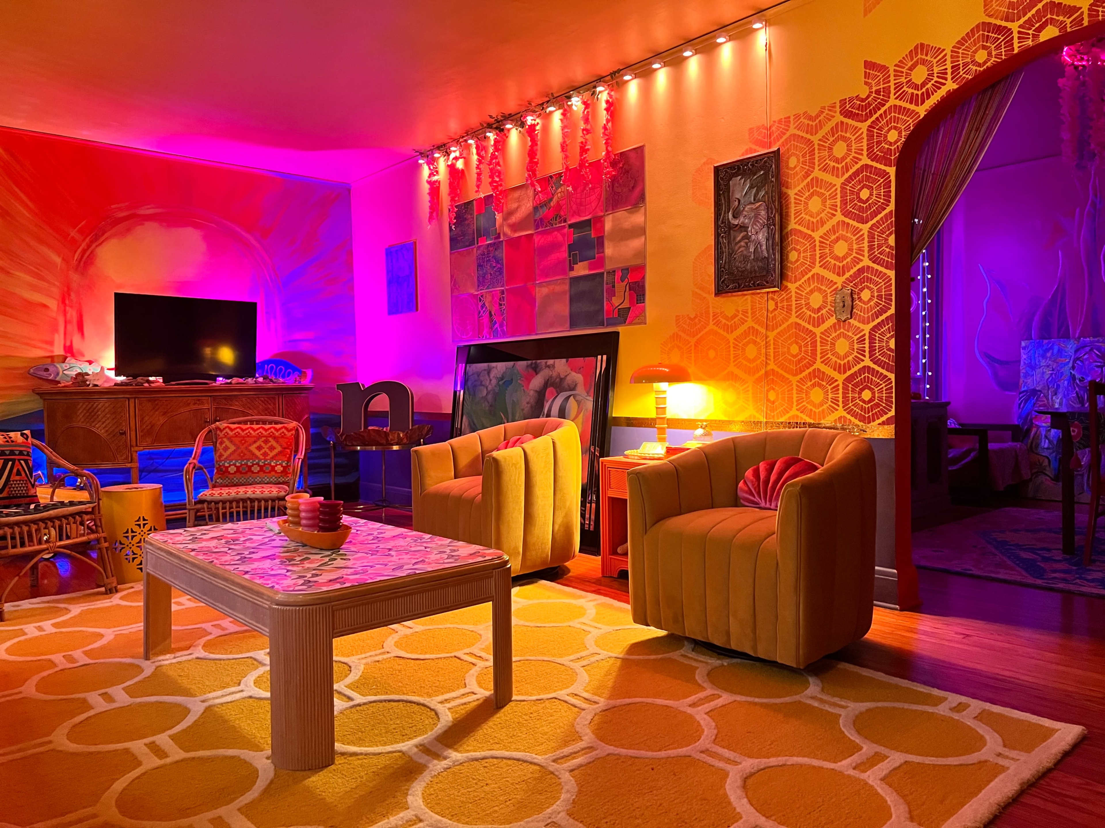 The room features colorful walls, a geometric rug, and two yellow armchairs positioned around a patterned coffee table under vibrant lighting.