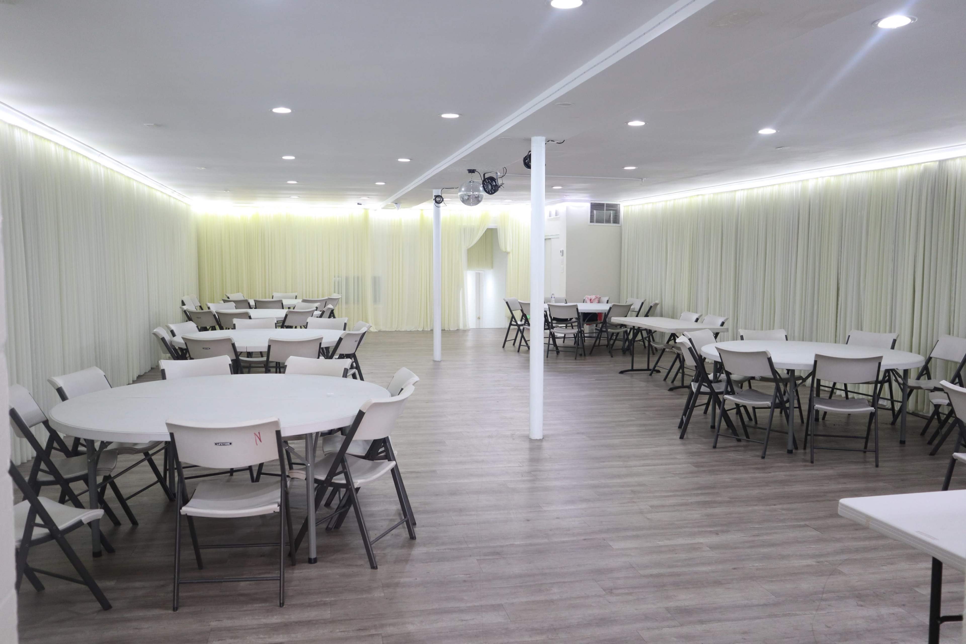 A large, empty event space with circular tables and folding chairs arranged throughout, illuminated by soft lights along the wall.