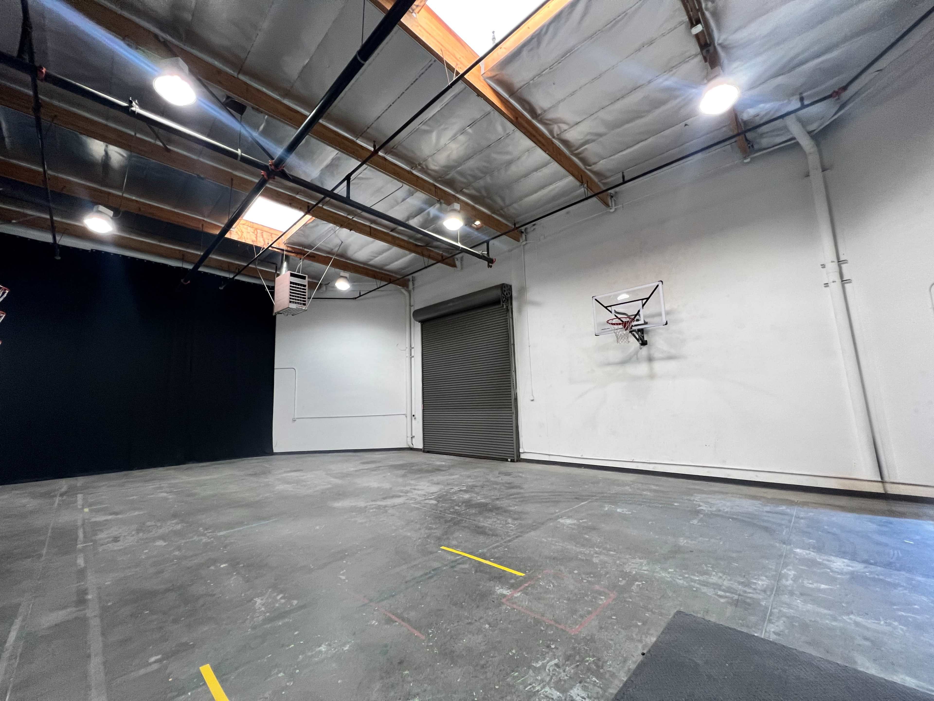 A spacious, empty warehouse interior with exposed beams, a basketball hoop mounted on the wall, and a large rolling door closed at one end.