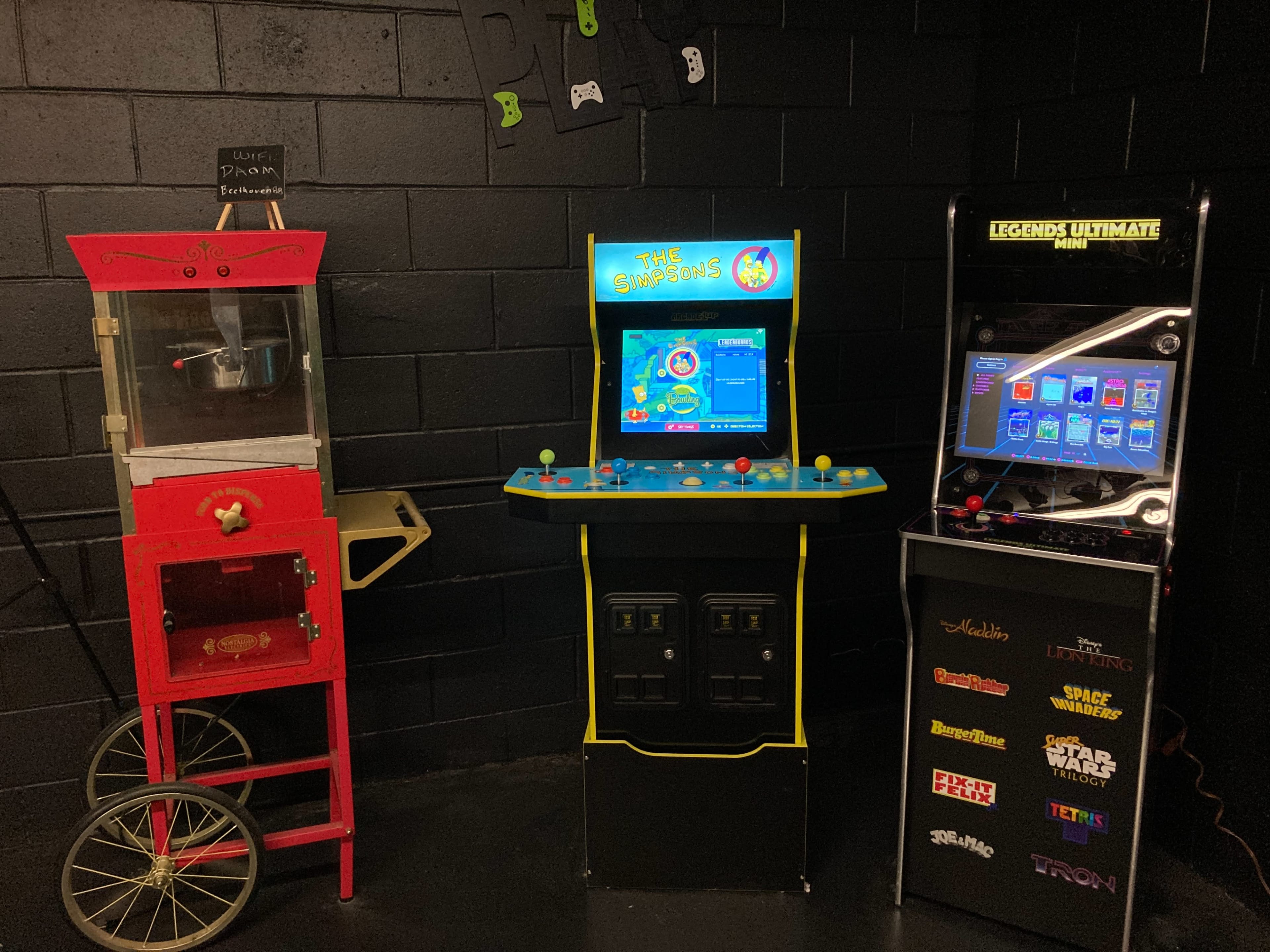 A game room with a vintage popcorn machine, an arcade console featuring "The Simpsons," and a multi-game arcade machine displaying a variety of titles on its screen.