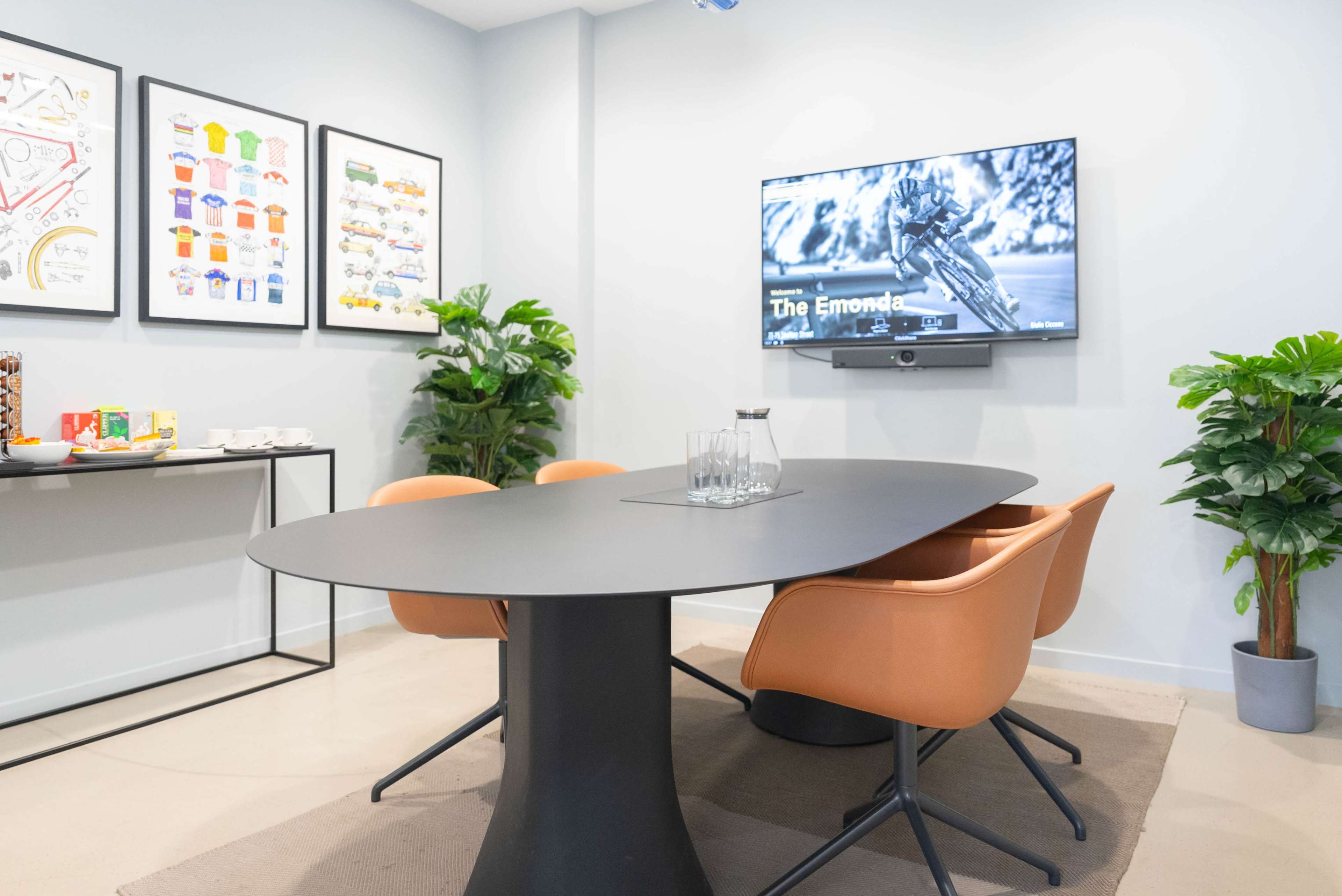 The image shows a modern conference room featuring a large oval table surrounded by four orange chairs, with a wall-mounted TV and decorative plants.