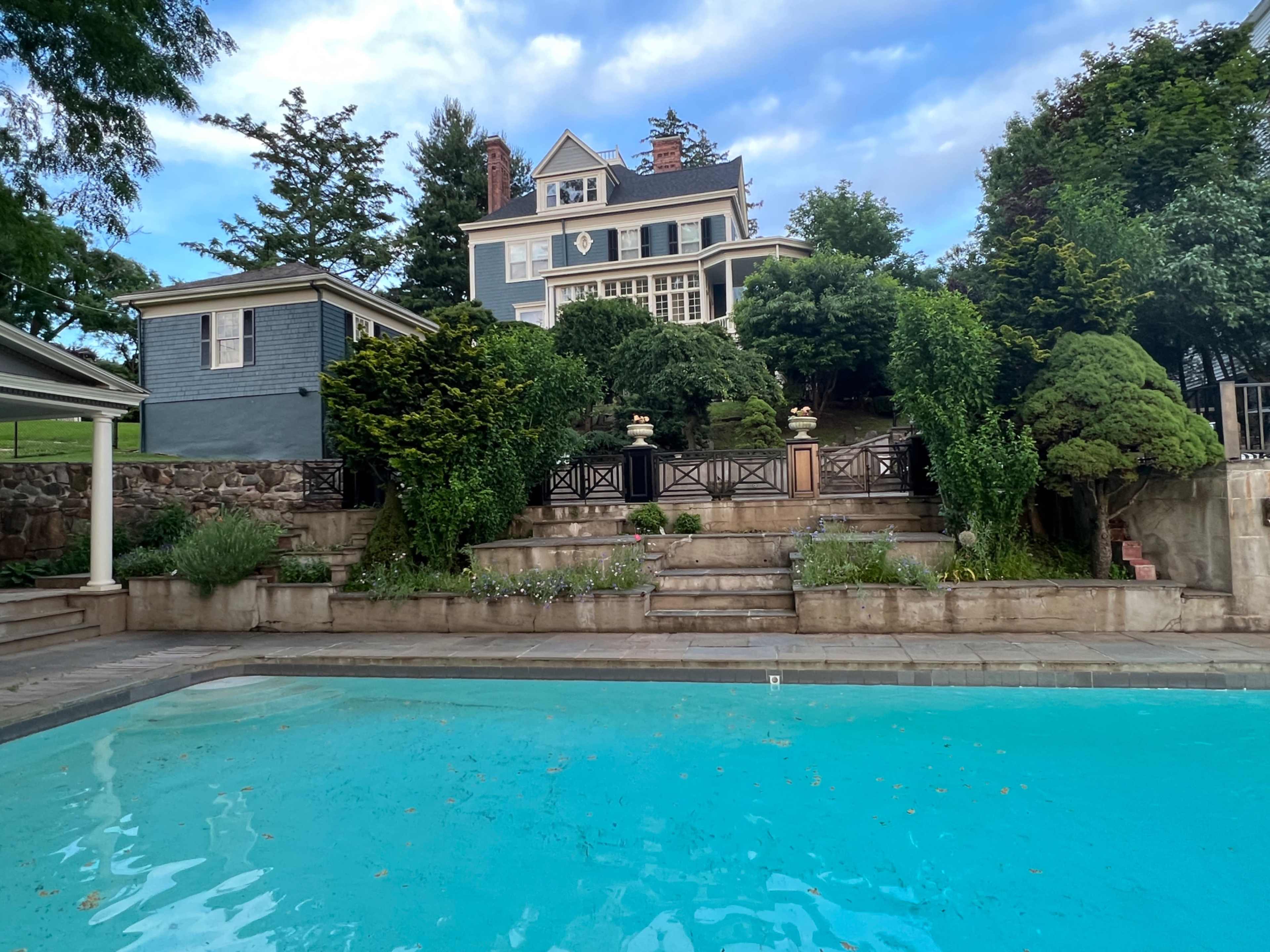 A large blue house sits on a hillside surrounded by greenery, overlooking a turquoise swimming pool in the foreground.