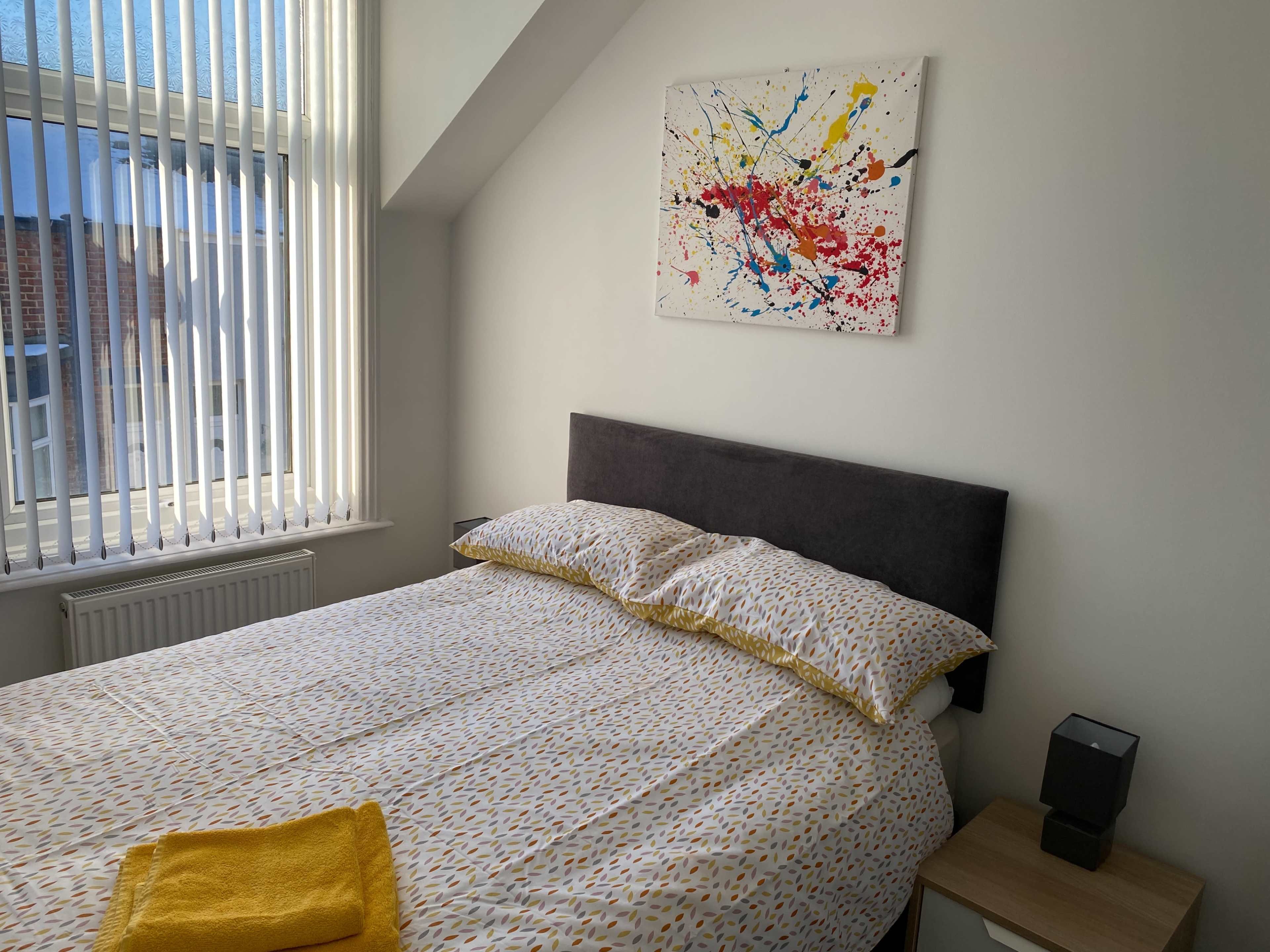 The image shows a neatly made bed in a bright room, with a colorful abstract painting above the headboard and a yellow towel folded at the foot of the bed.