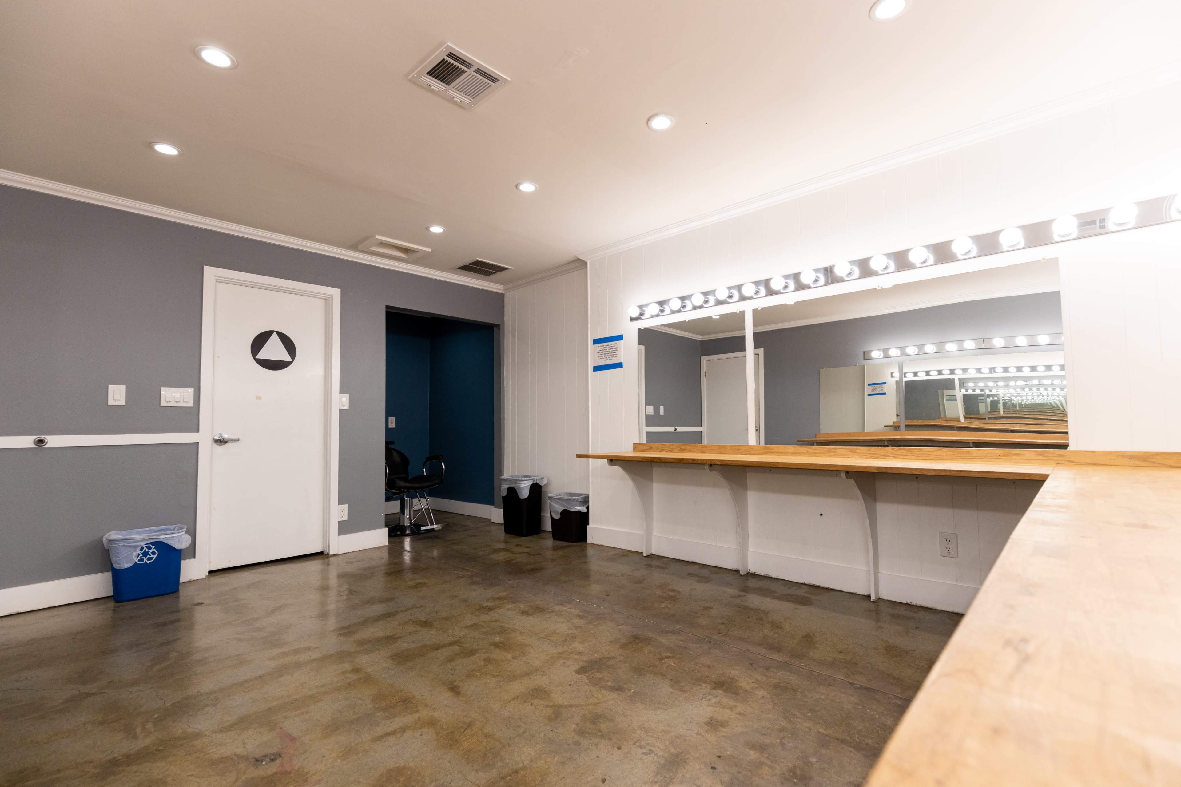 The image shows a spacious, well-lit room with a wooden counter, a large mirror with bulbs, and minimal furniture, including a chair and waste bins.