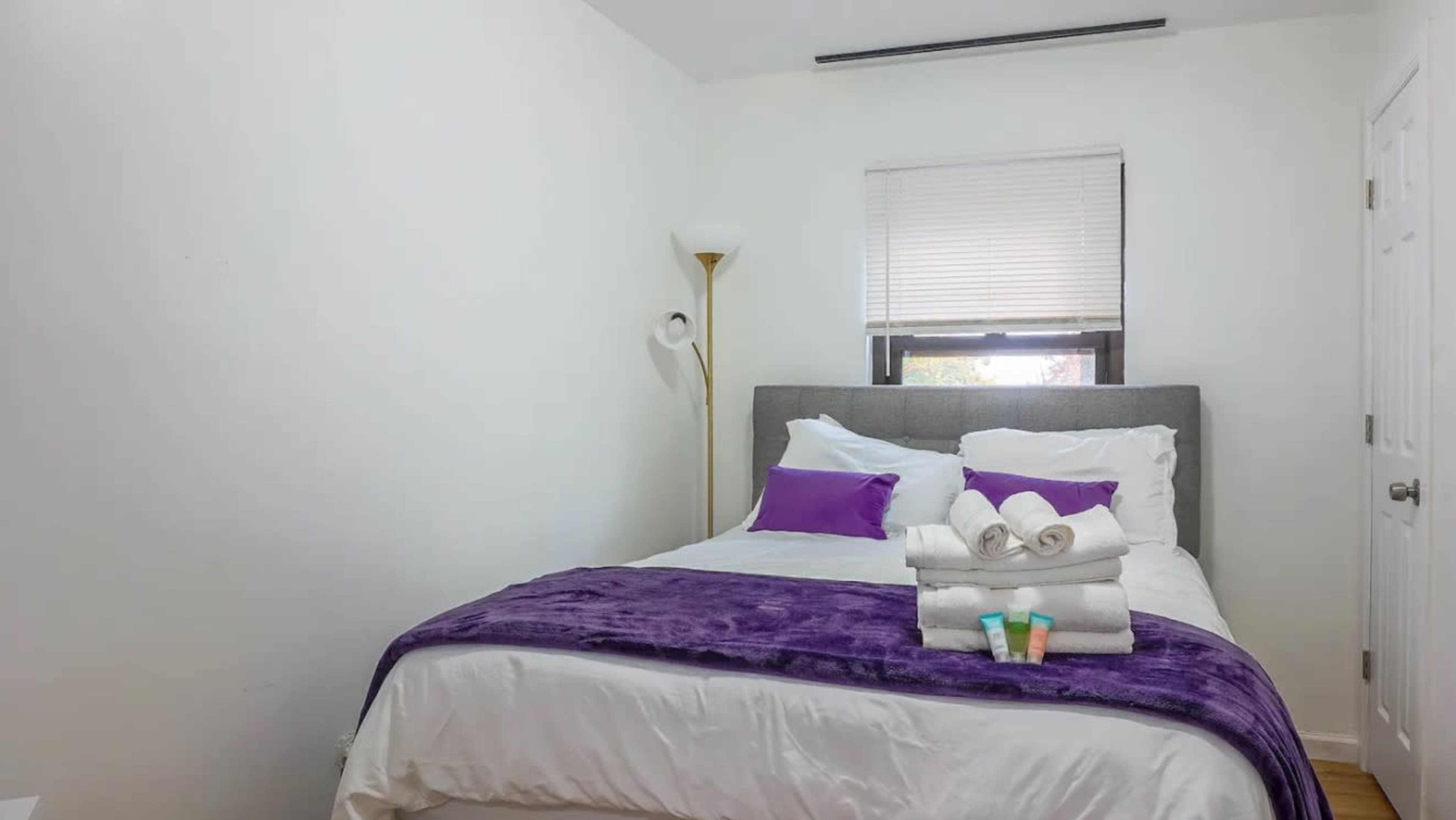 A neatly arranged bedroom features a bed with white linens and purple accents, along with towels and toiletries neatly placed on the bed.