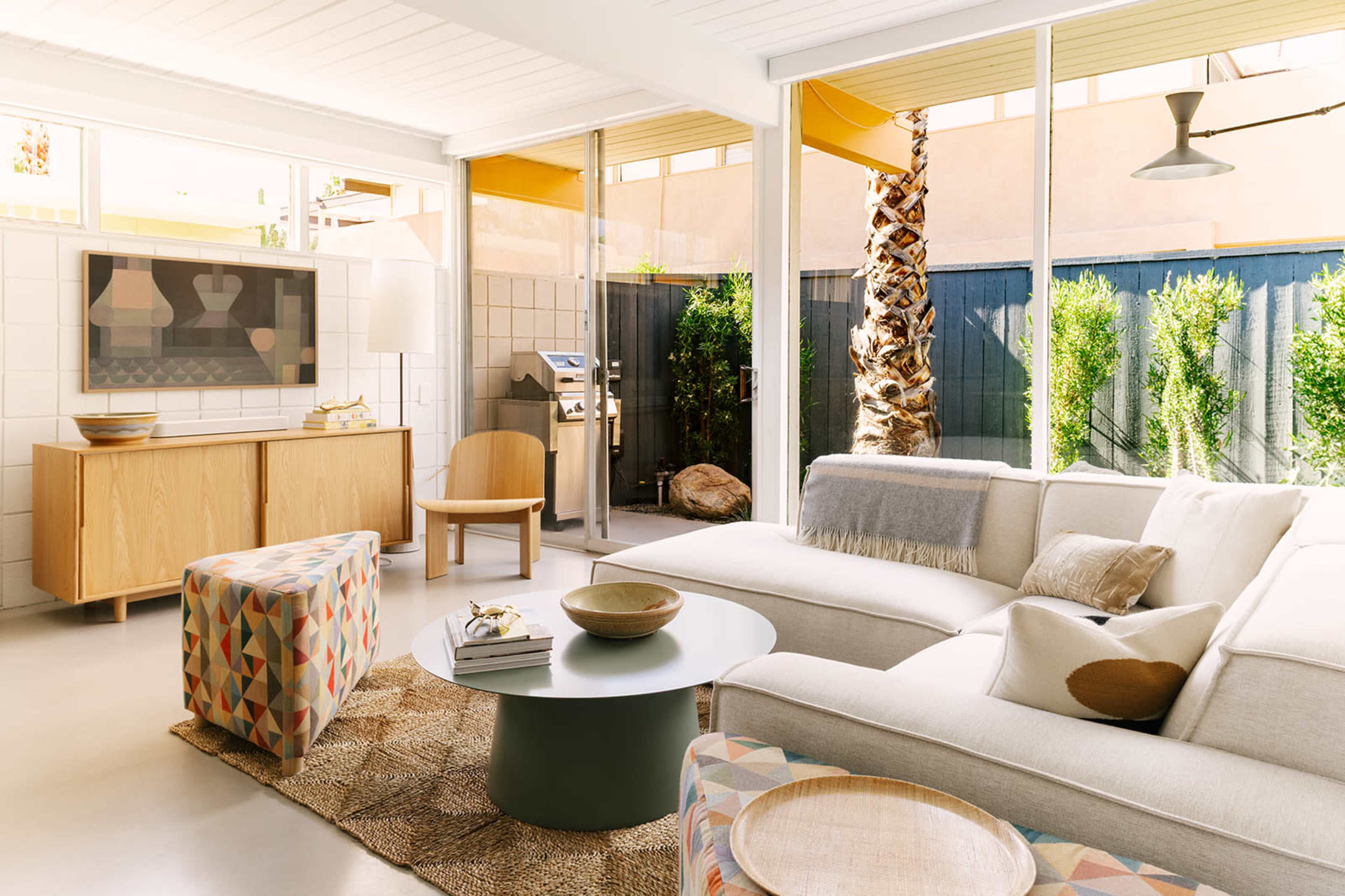 The image shows a modern living room with a light-colored sectional sofa, a round coffee table, and a view of a patio with greenery and a grill outside.