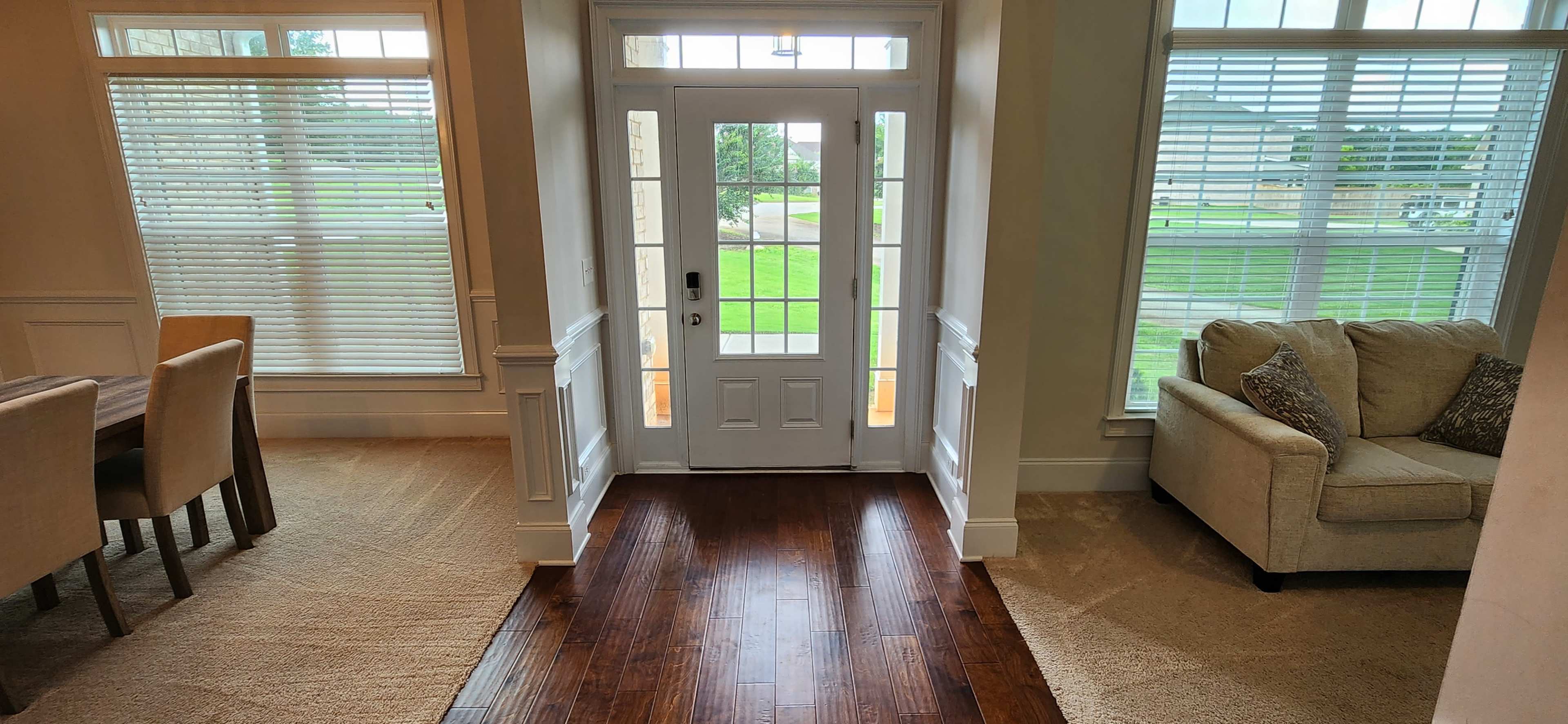 A hallway leading to a front door, with a large window on one side and a dining area with a table on the other, featuring wooden flooring and neutral-colored furnishings.
