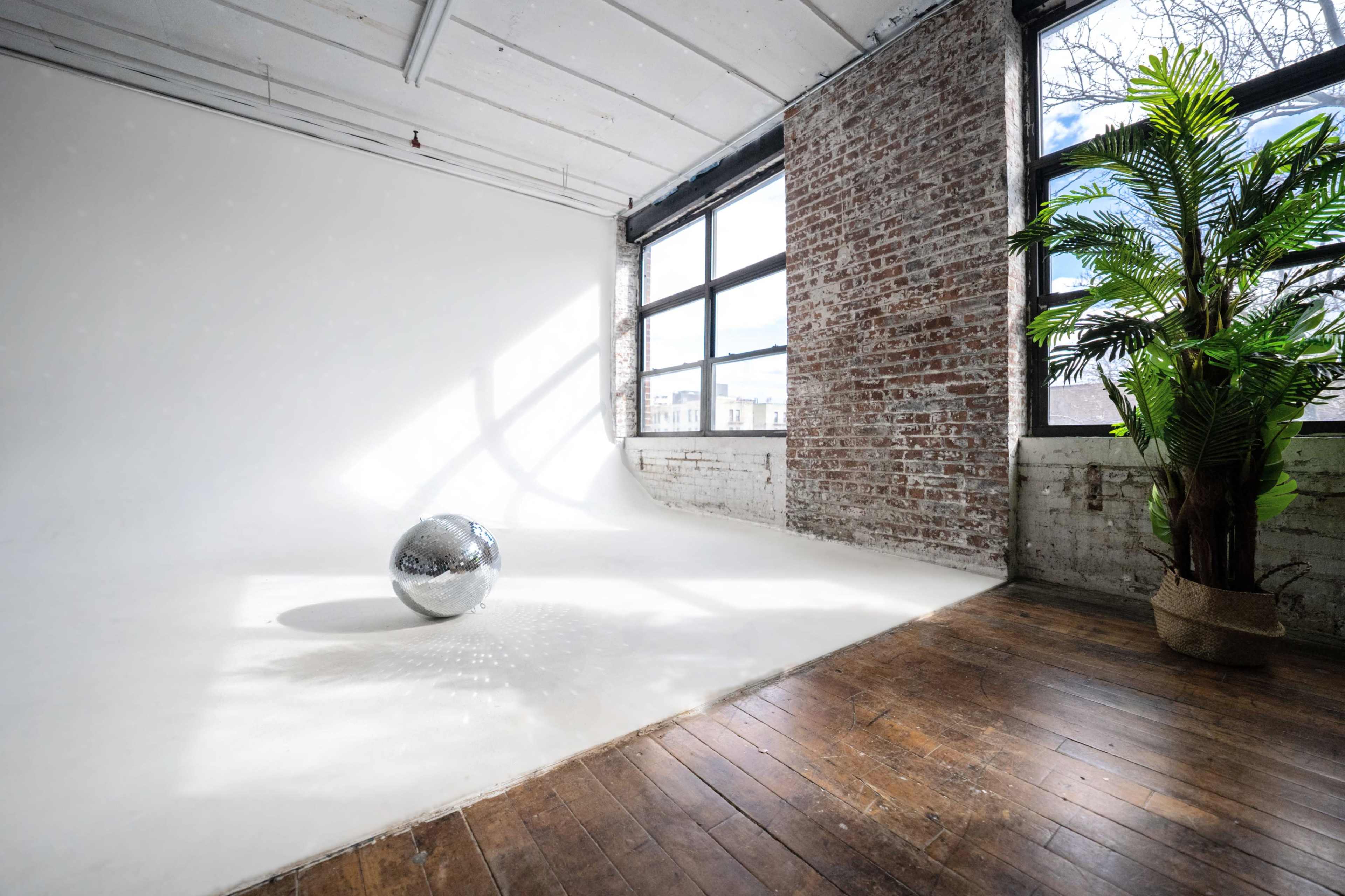 A mirrored disco ball sits on a white curved backdrop in a spacious room with large windows and a potted plant.