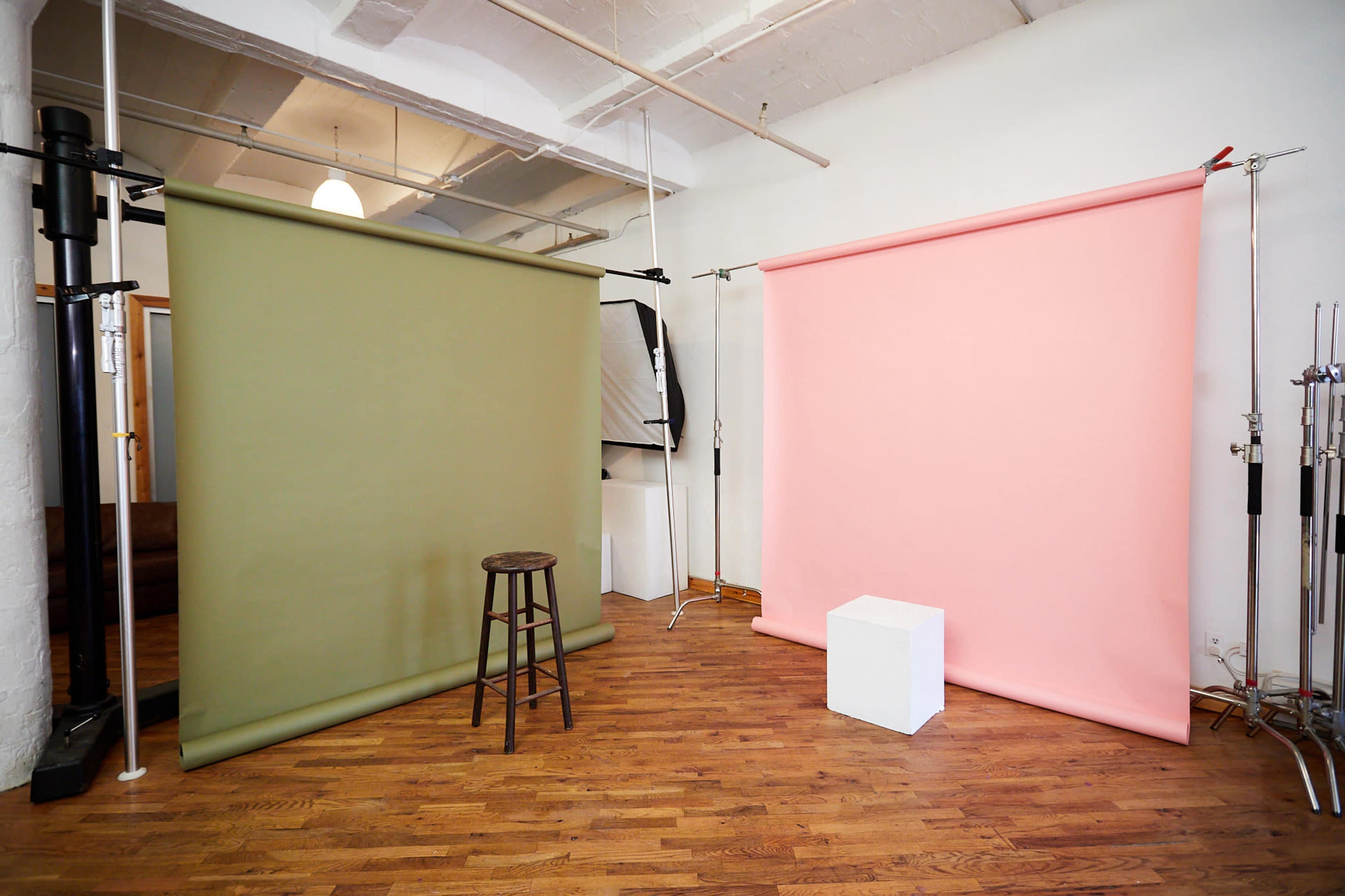 A photography studio features green and pink backdrops on either side, with a wooden stool and a white cube in the center.