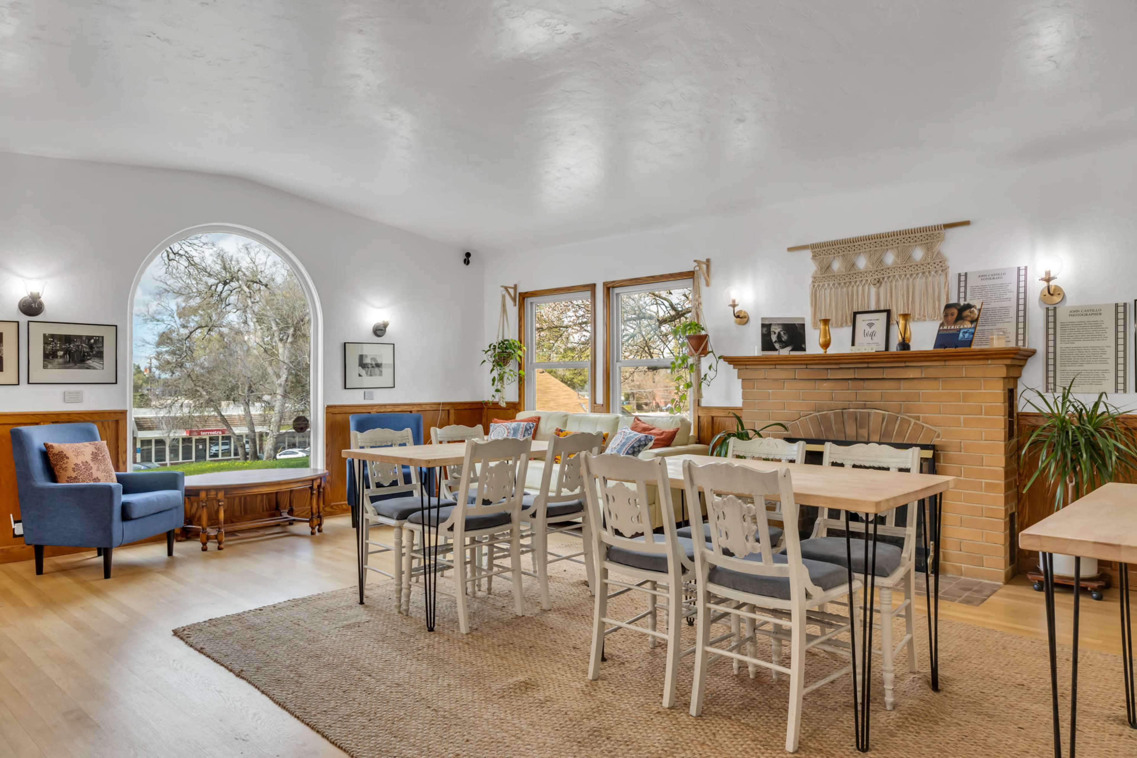 A spacious living area featuring a blue armchair, a large window, a dining table with white chairs, and a brick fireplace.