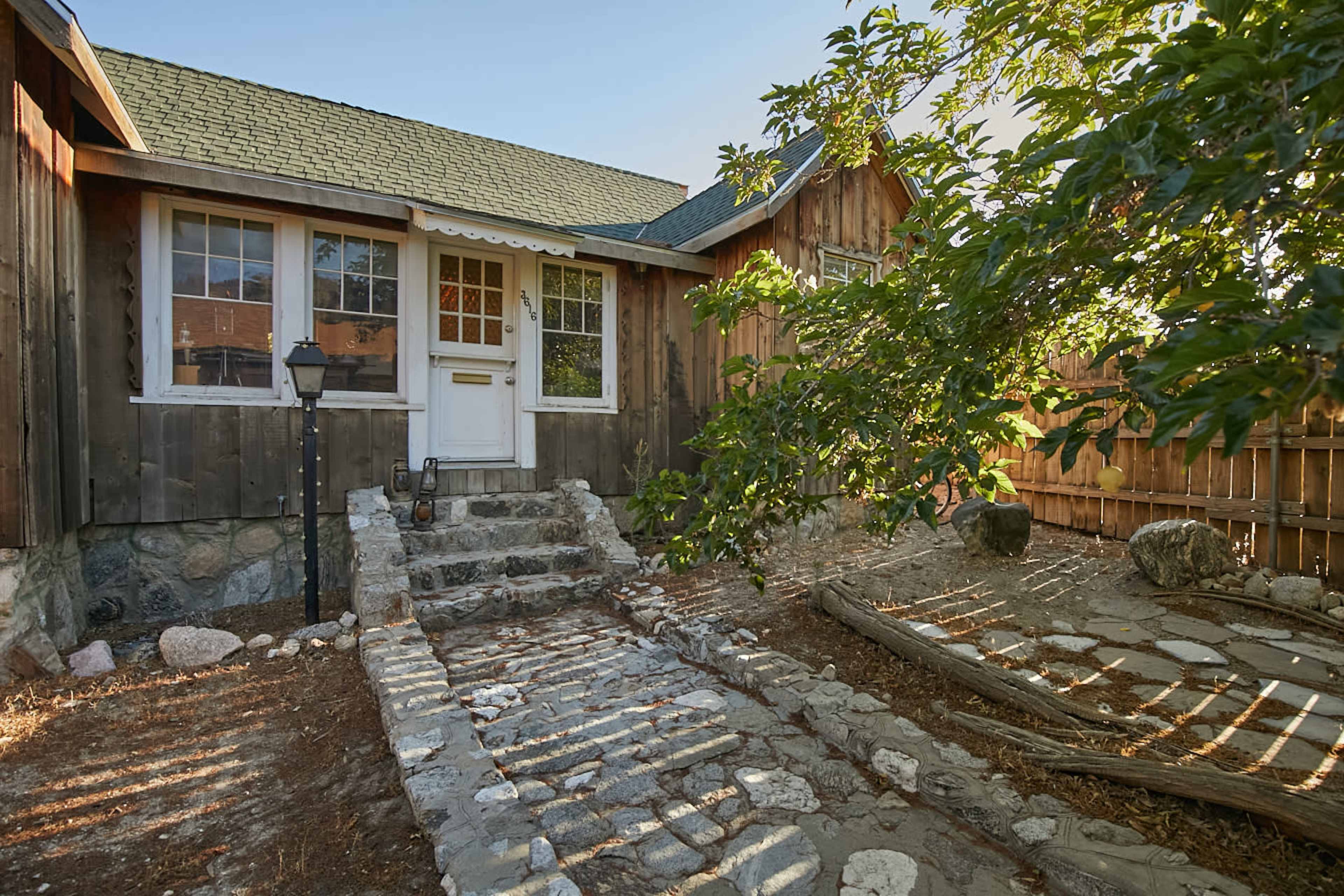 A stone pathway leads to the front door of a wooden house, surrounded by a fence and greenery.