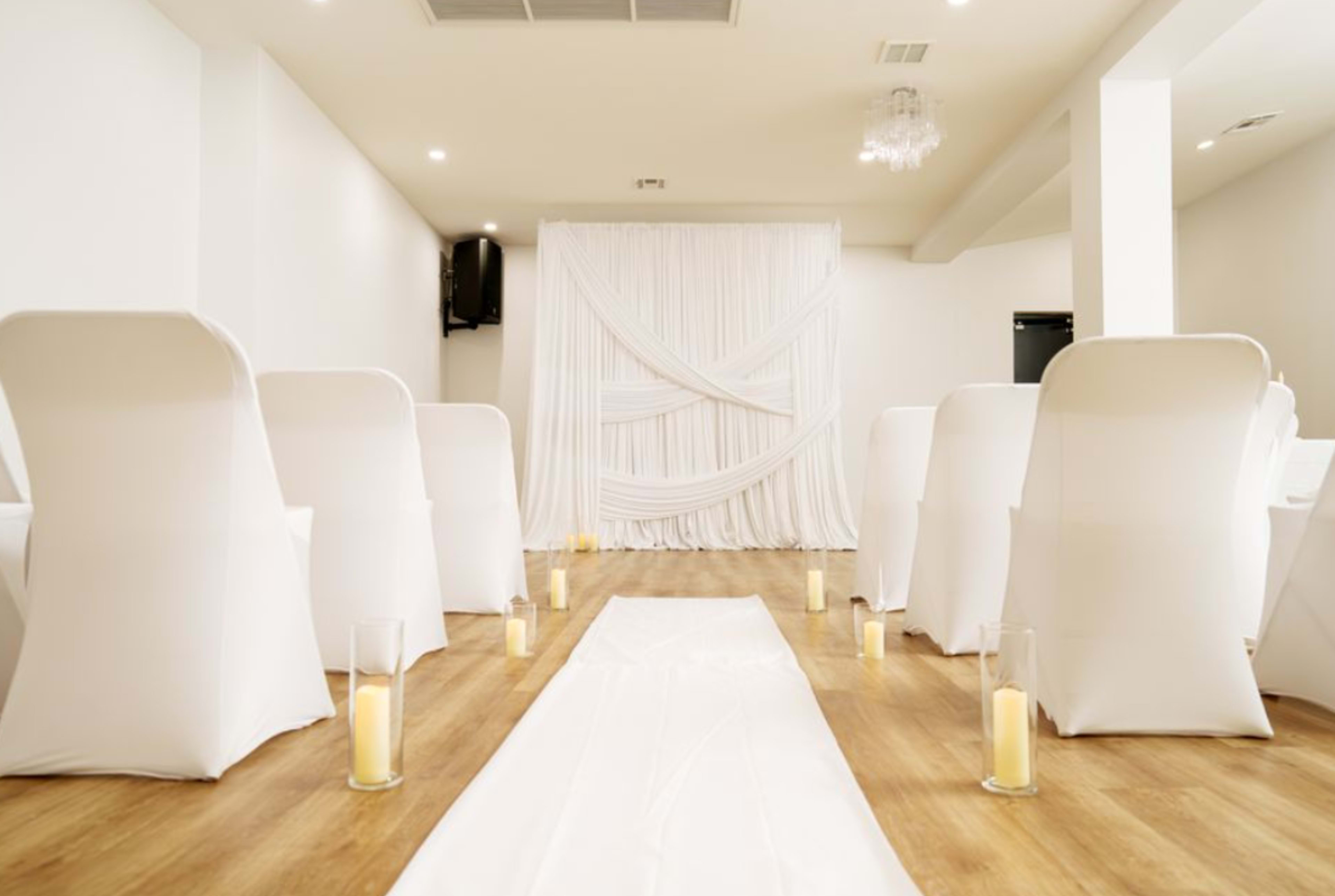 A minimalist event space is set up with rows of white chairs facing a draped backdrop, complemented by candles arranged along a central white aisle runner.