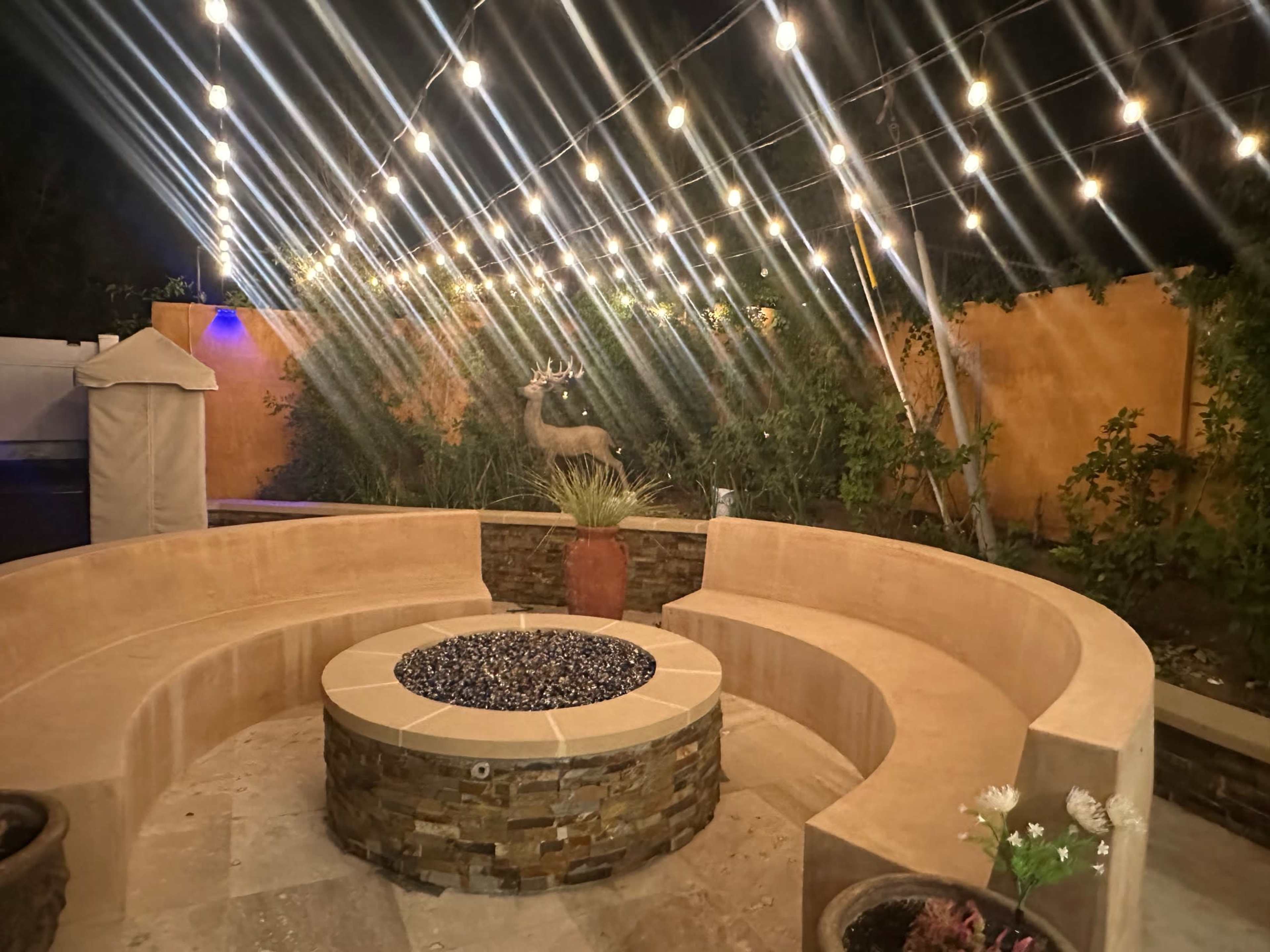 The image shows a circular stone seating area with a fire pit in the center, surrounded by plants and illuminated by string lights overhead.