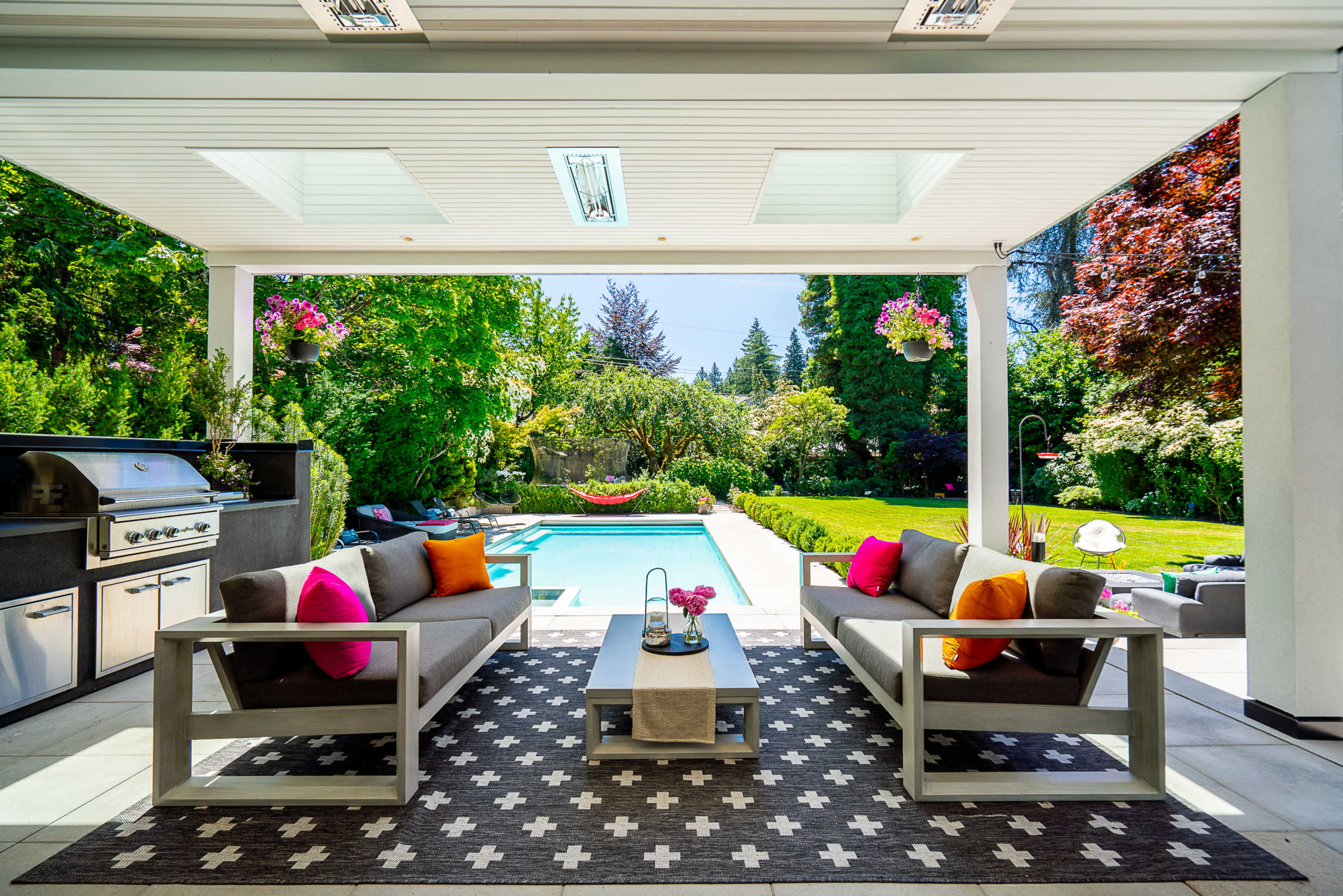 A covered patio features two gray sofas adorned with colorful pillows, facing a small coffee table overlooking a landscaped garden with a swimming pool.