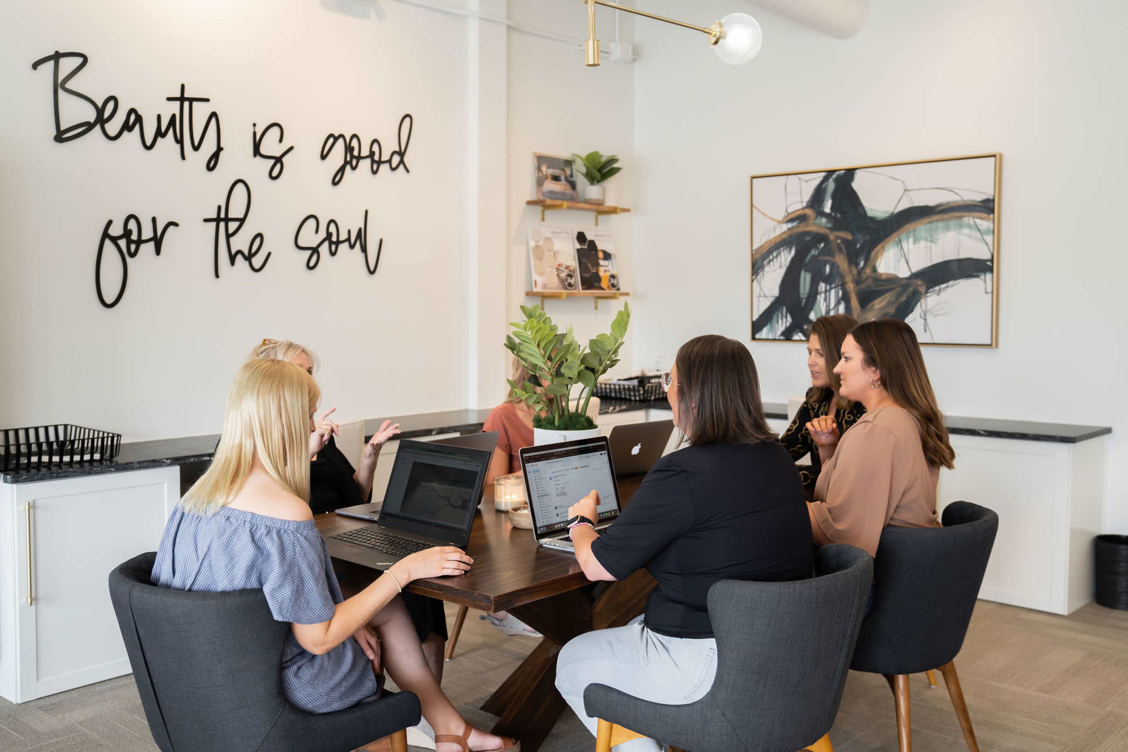 A group of six people are gathered around a table with laptops in a well-lit meeting room featuring a decorative wall with the phrase "Beauty is good for the soul."