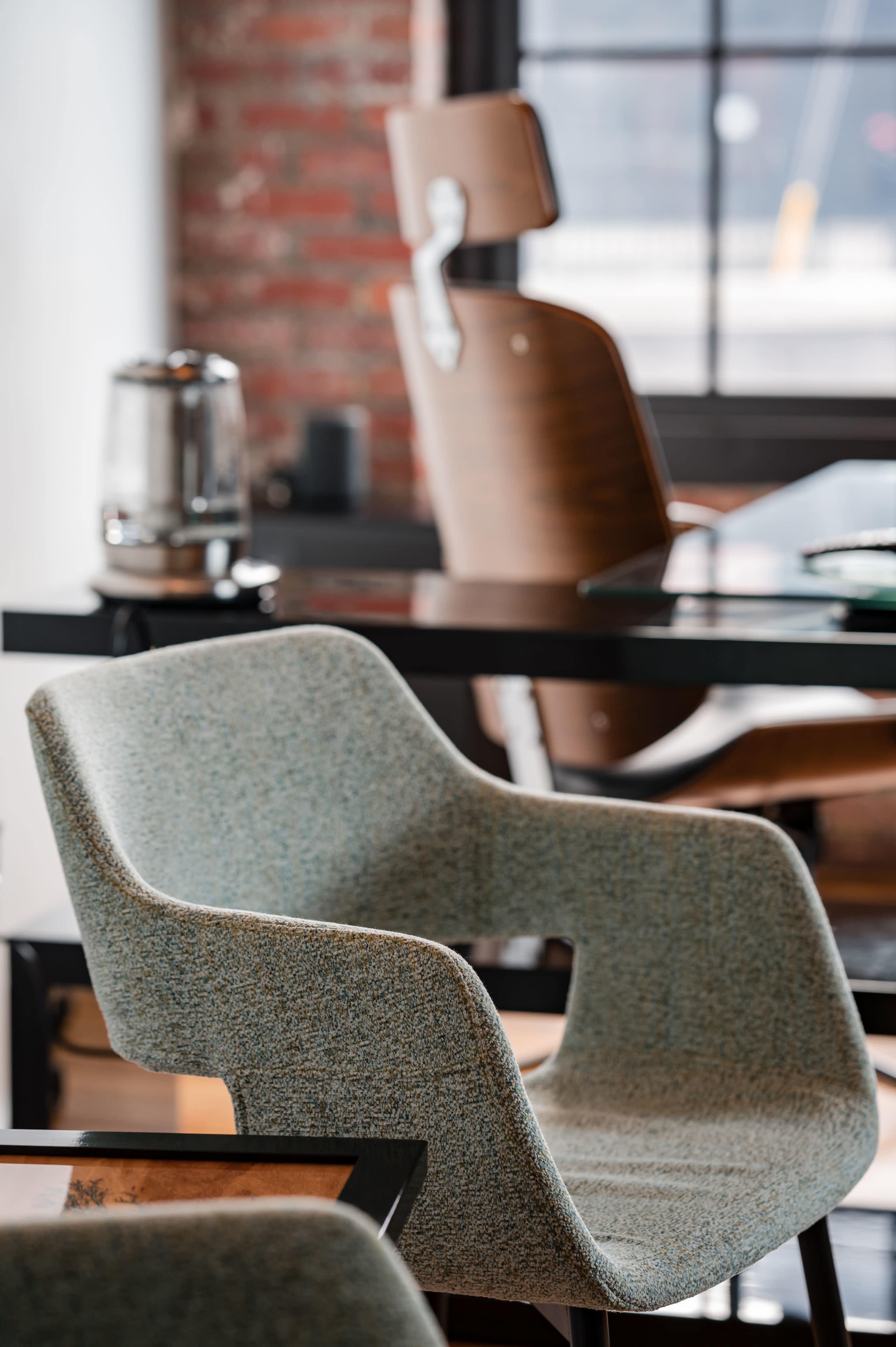 A modern workspace featuring a patterned chair in the foreground, with a wooden chair and a glass table in the background against a brick wall.