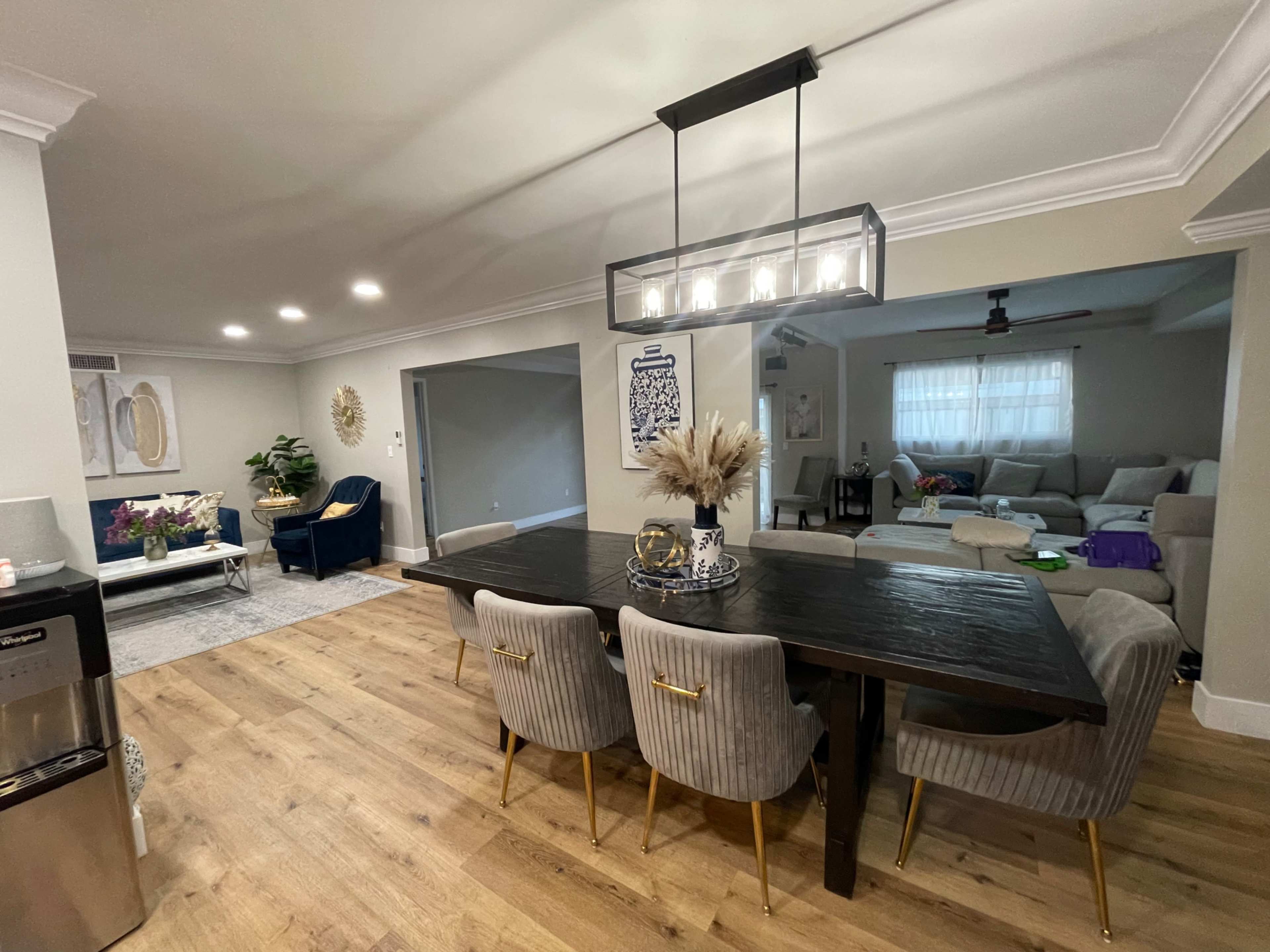 A modern dining area with a large wooden table surrounded by upholstered chairs, leading to a living room with a sectional sofa and a large window.