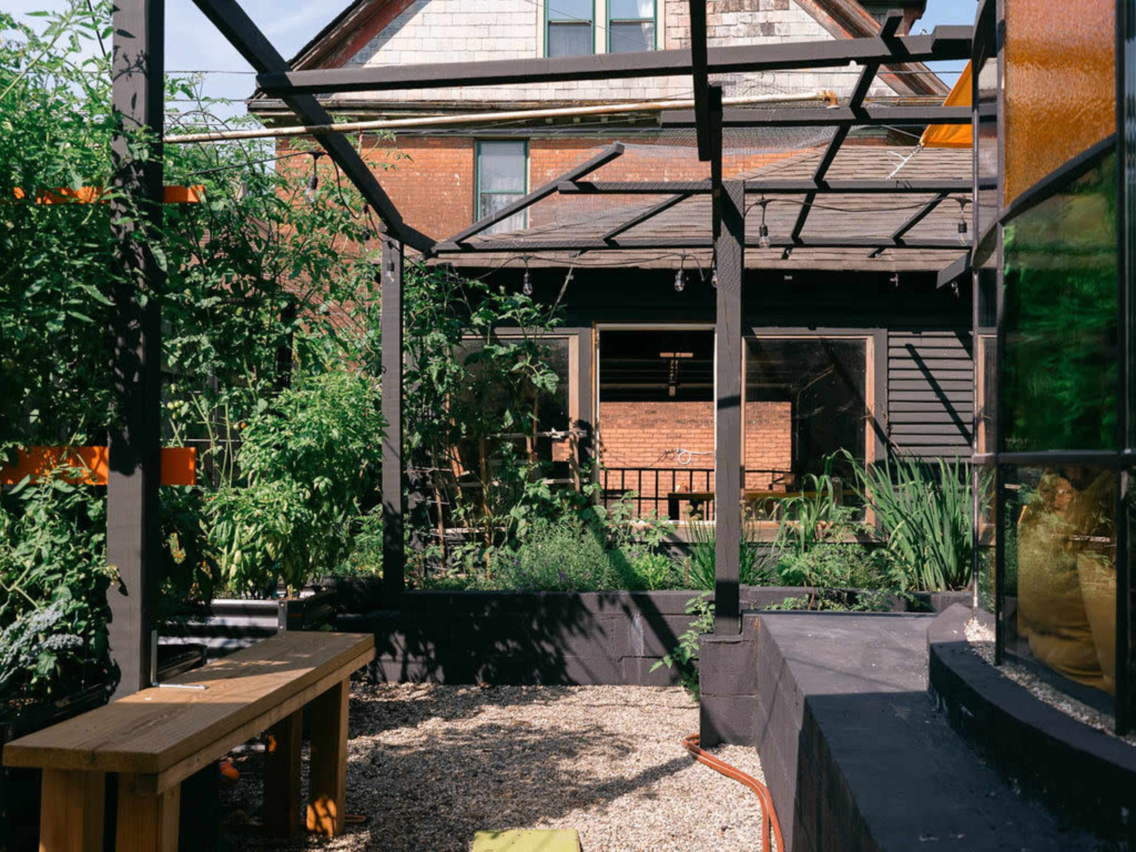 The image shows a backyard garden with raised planters filled with plants, a wooden bench, and a view of a house with large windows.