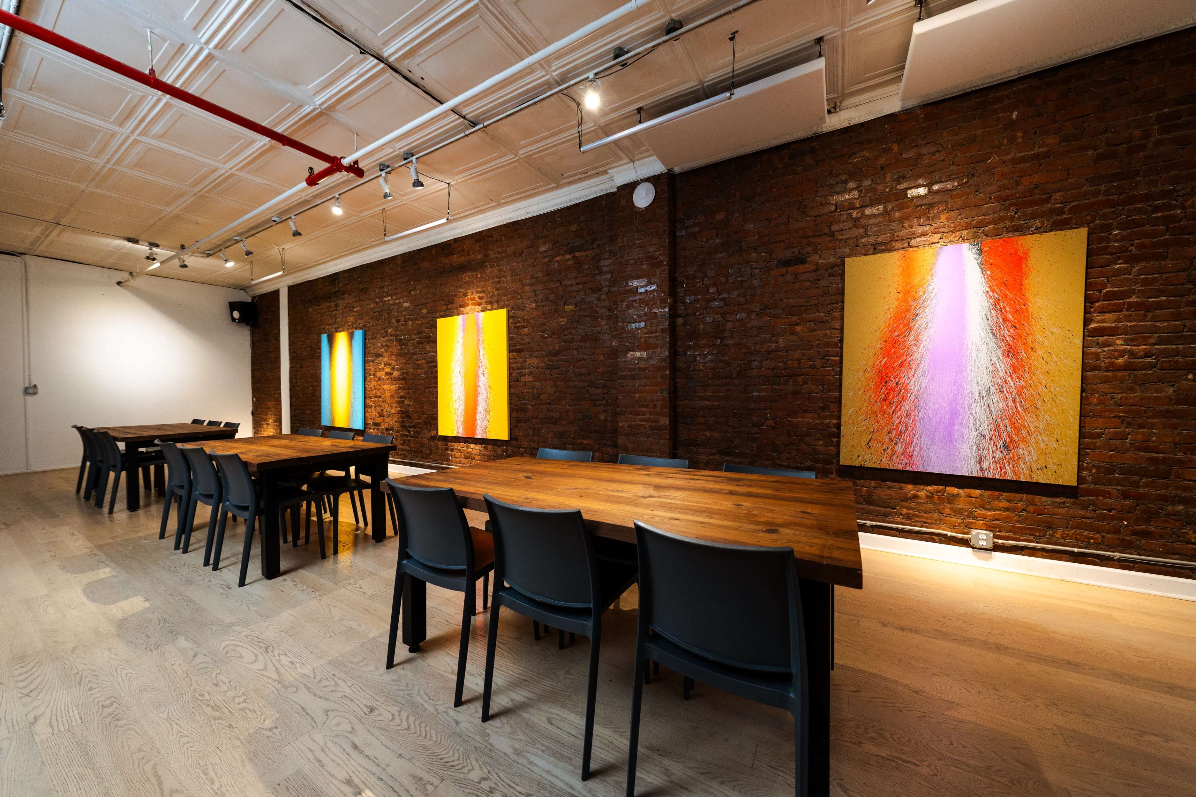 The image shows a modern meeting room with a long wooden table, black chairs, and colorful artwork on the exposed brick walls.