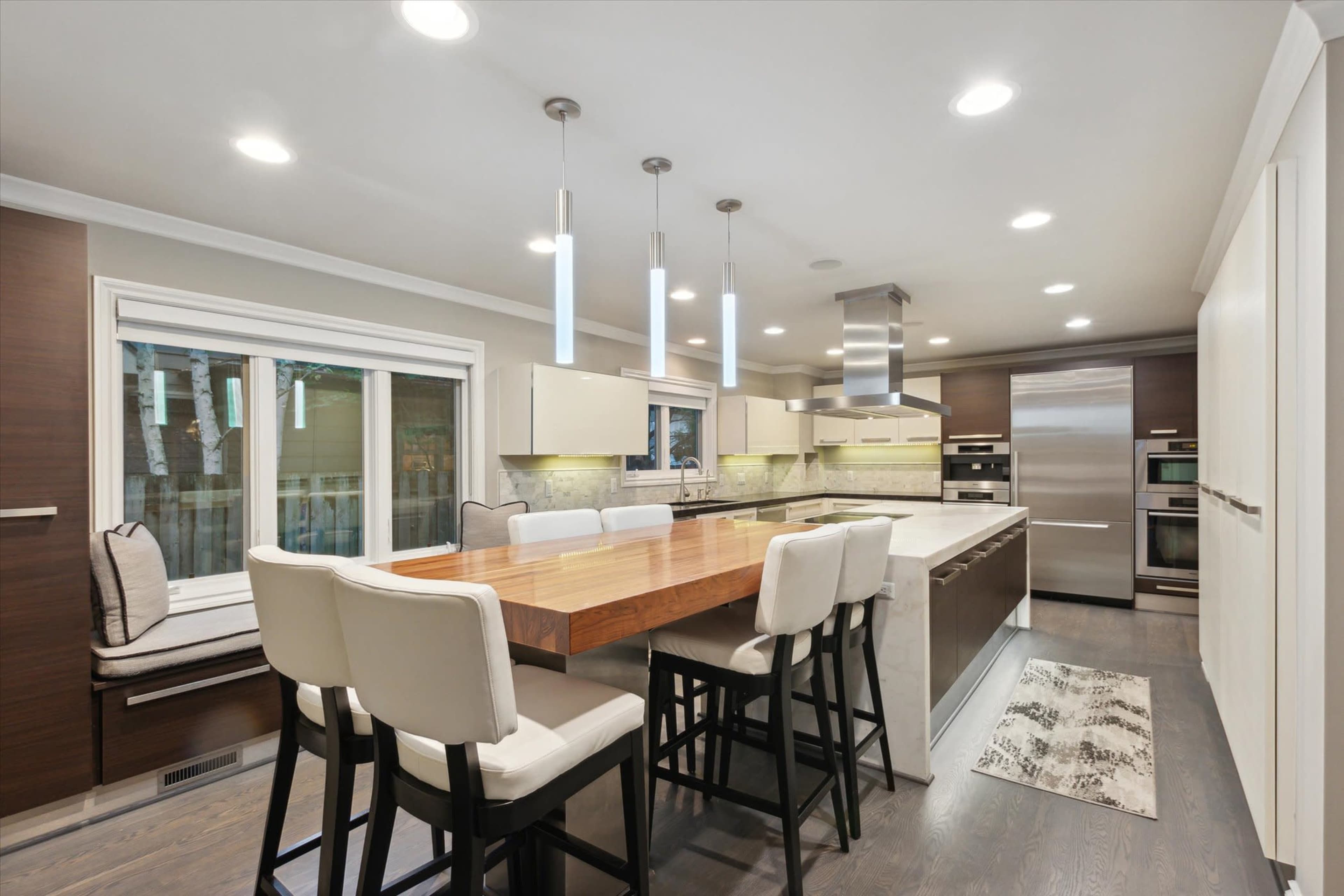A modern kitchen features a large wood dining table with high-backed chairs, stainless steel appliances, and ample natural light from a side window.