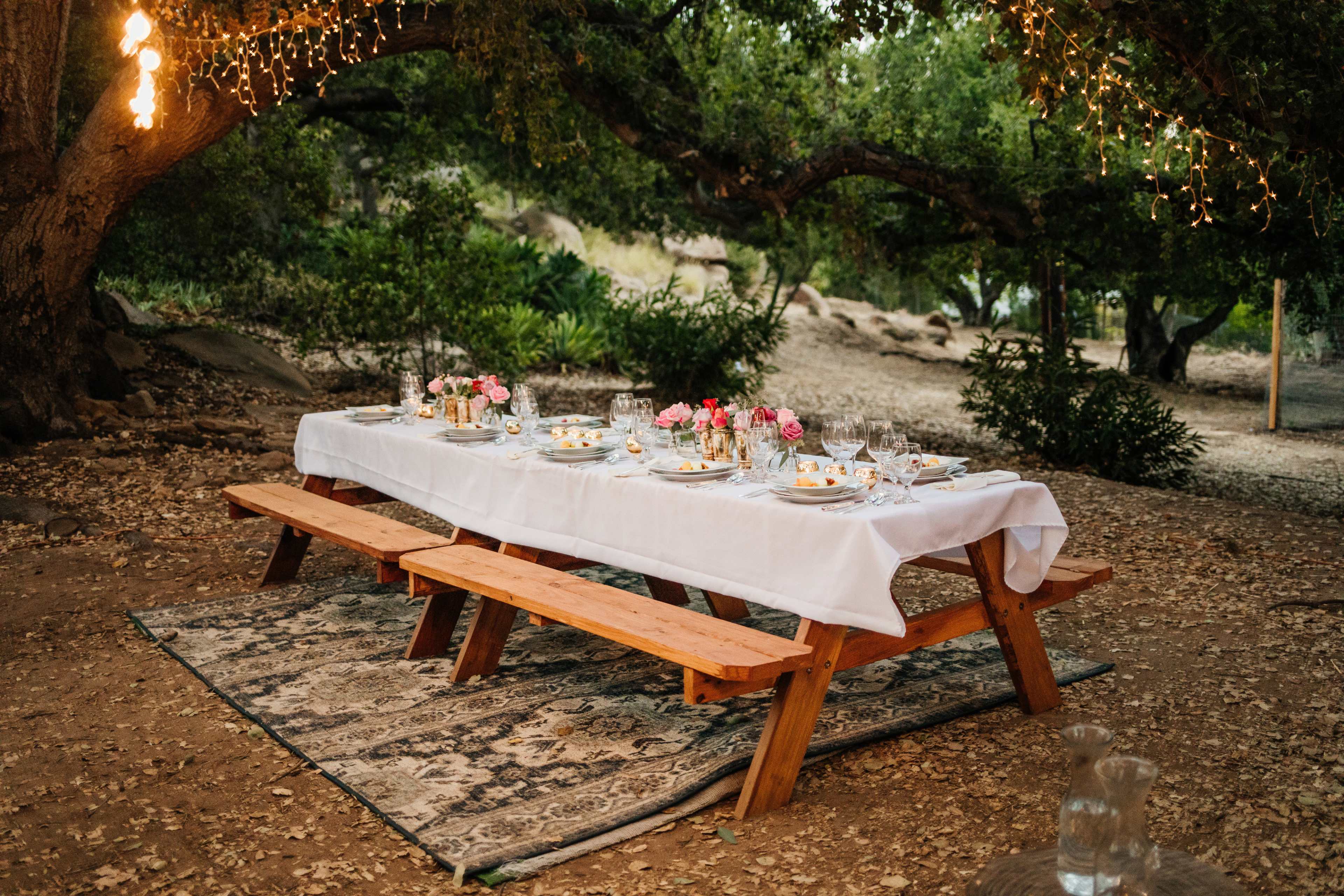 A long outdoor dining table is set with white tablecloths and adorned with flowers, surrounded by lush greenery and soft lighting.