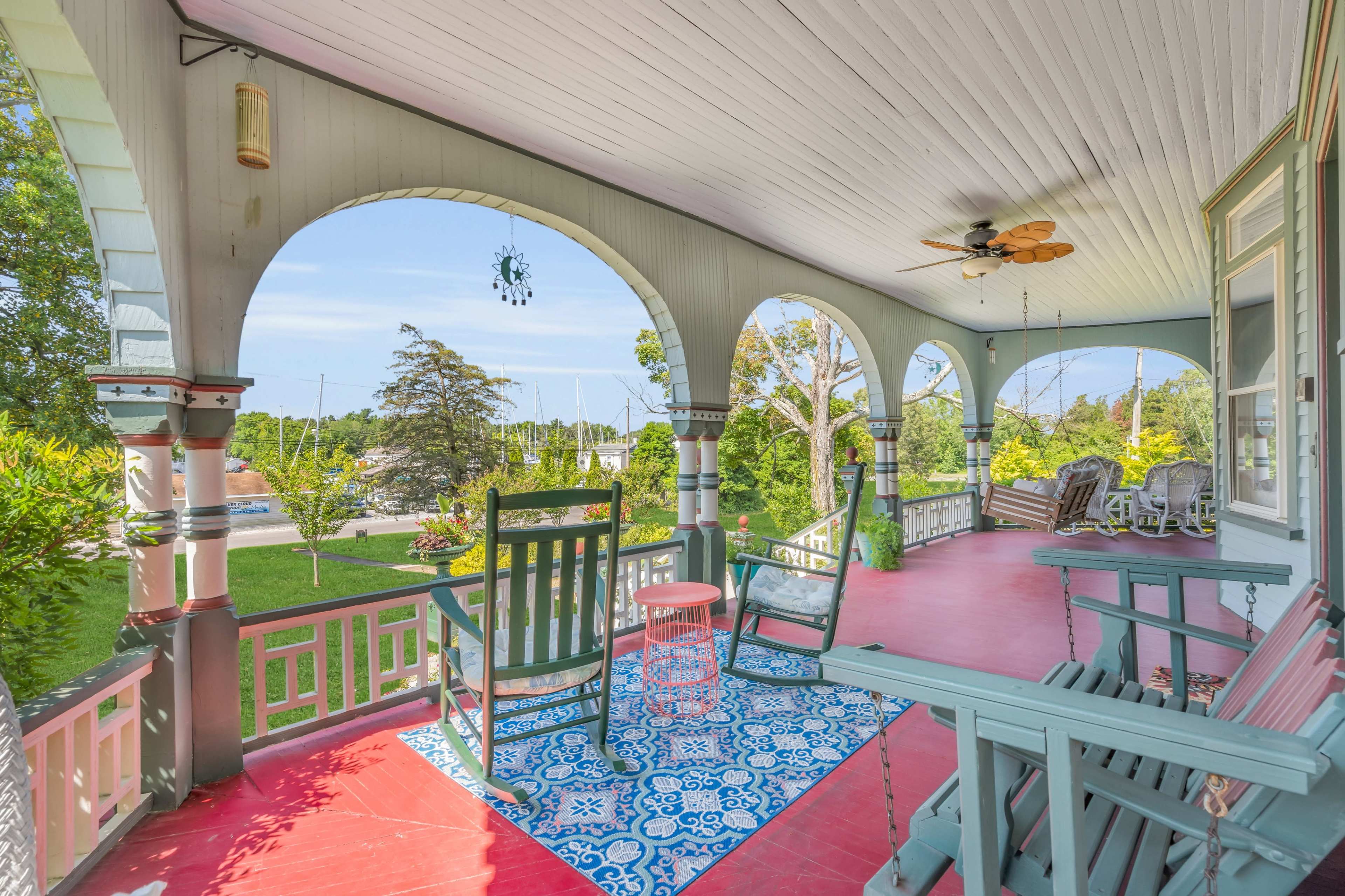 A spacious porch with arched openings, featuring rocking chairs, a small table, and a patterned rug, surrounded by greenery and visible boats in the distance.